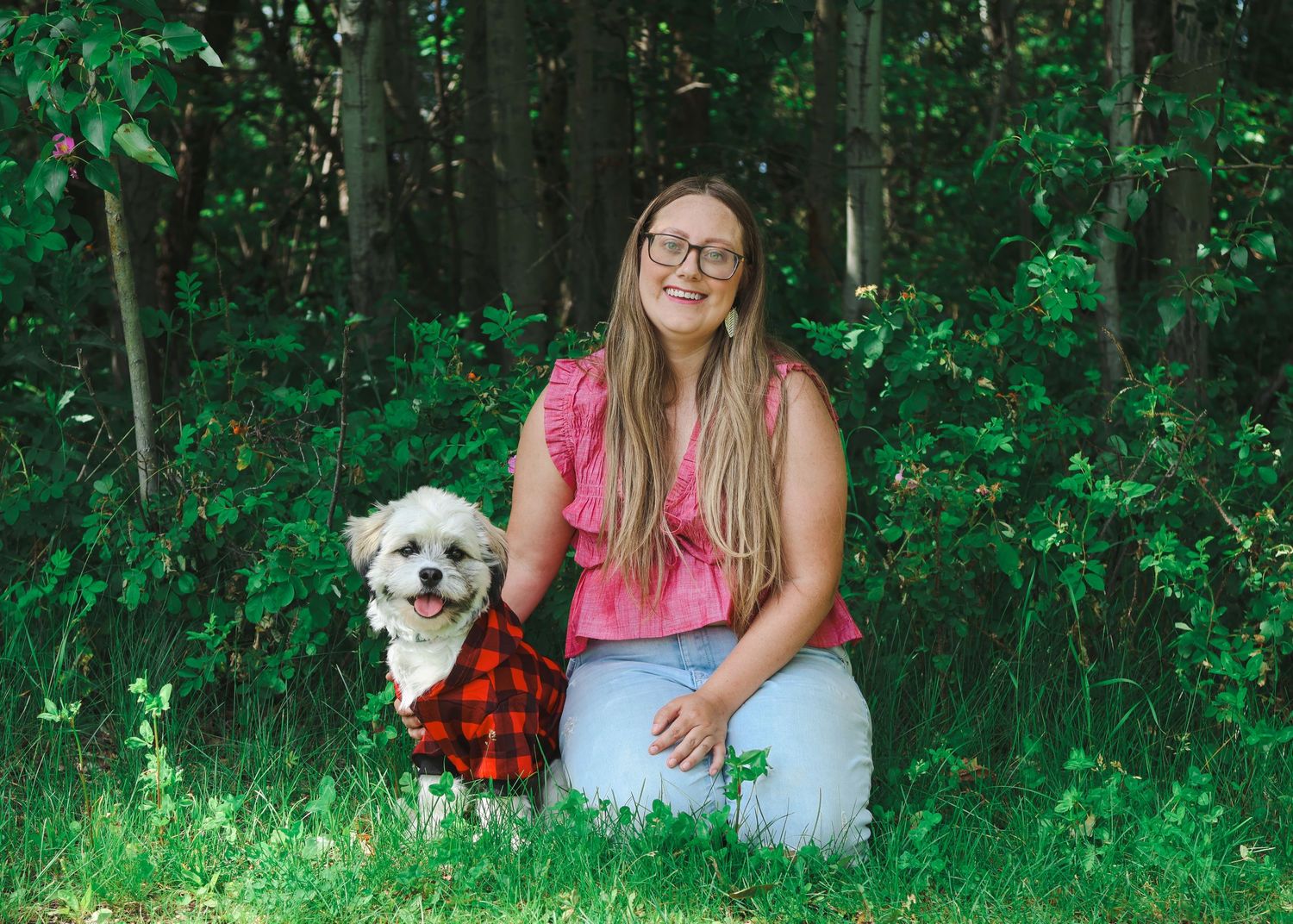 Woman with long blonde hair kneeling beside a small white dog in red plaid in a lush green setting.