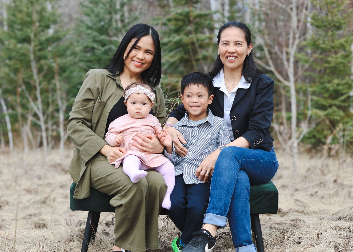 Two women pose outdoors with a baby and young boy in a forest setting during spring.