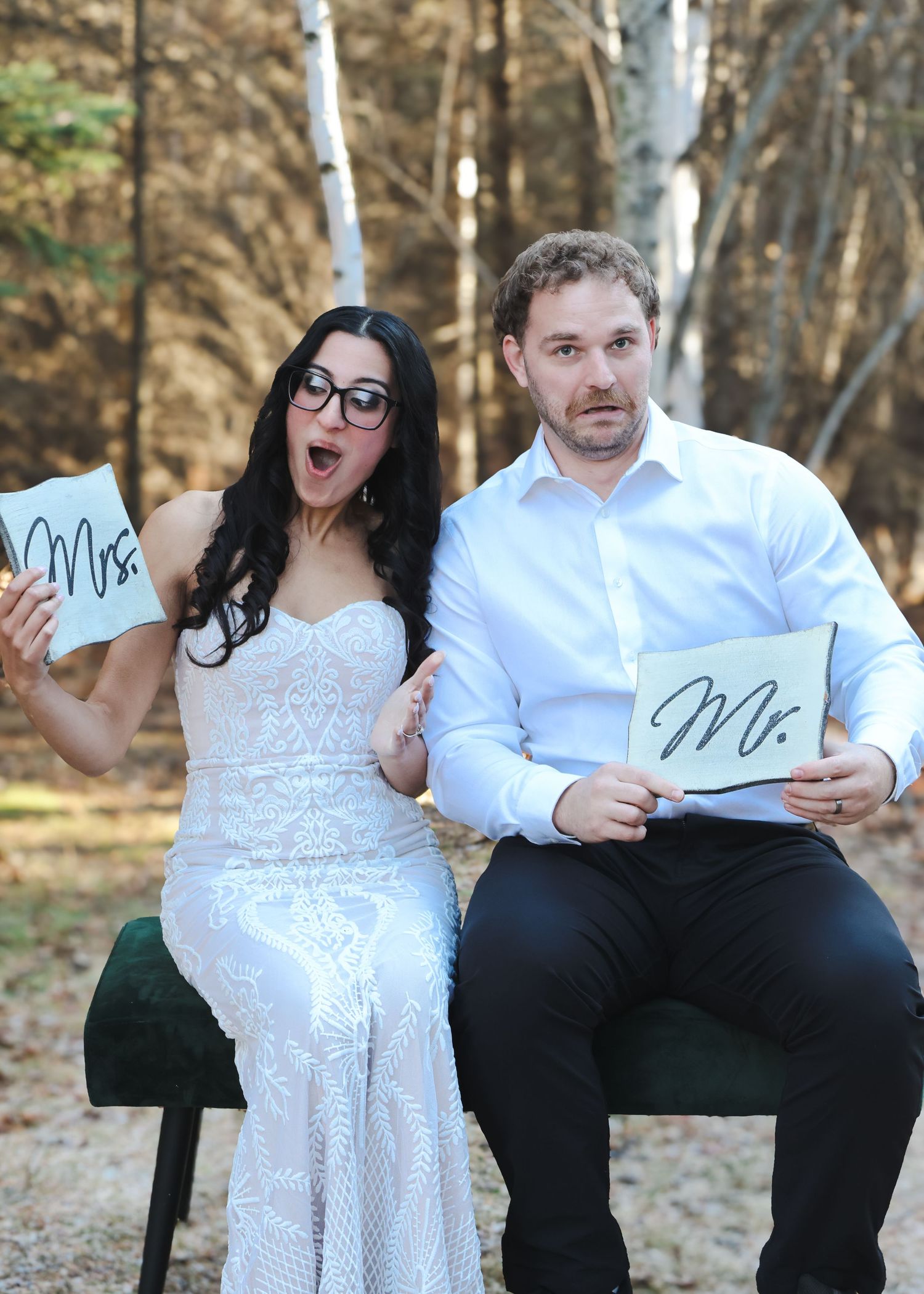 Playful bride and groom holding Mr. and Mrs. signs seated in autumn forest backdrop.