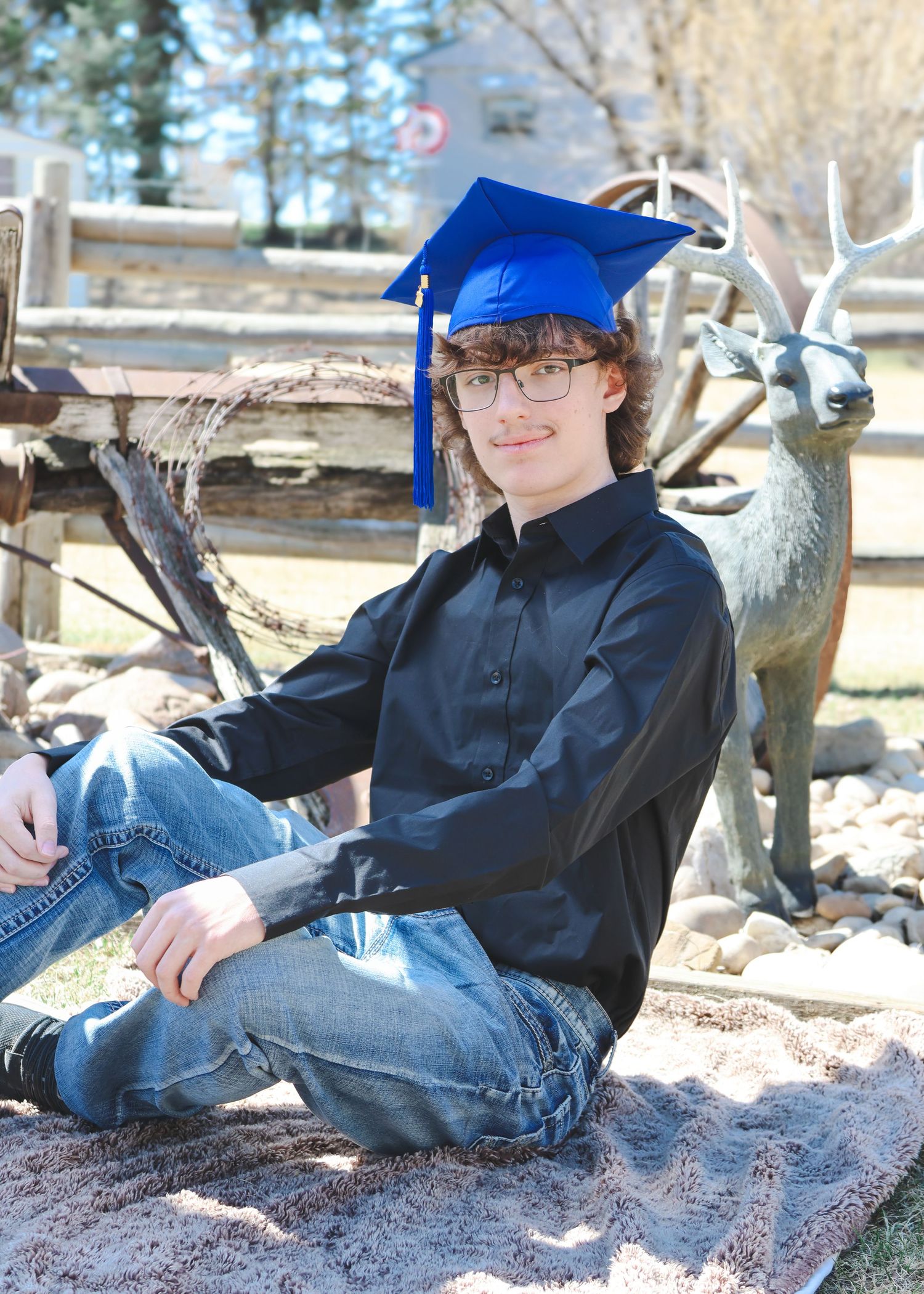 Male high school senior in blue graduation cap sitting outdoors near decorative deer statue.