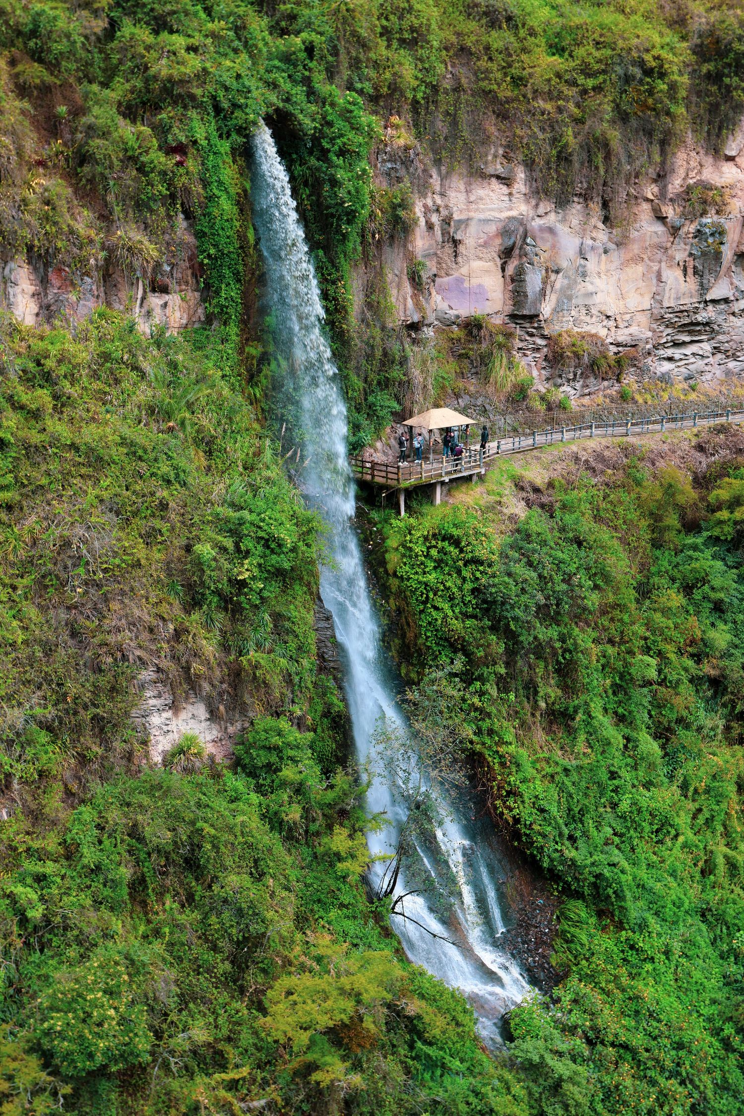 A tall waterfall cascades down a lush green cliff face with a wooden viewing platform built into the rock.