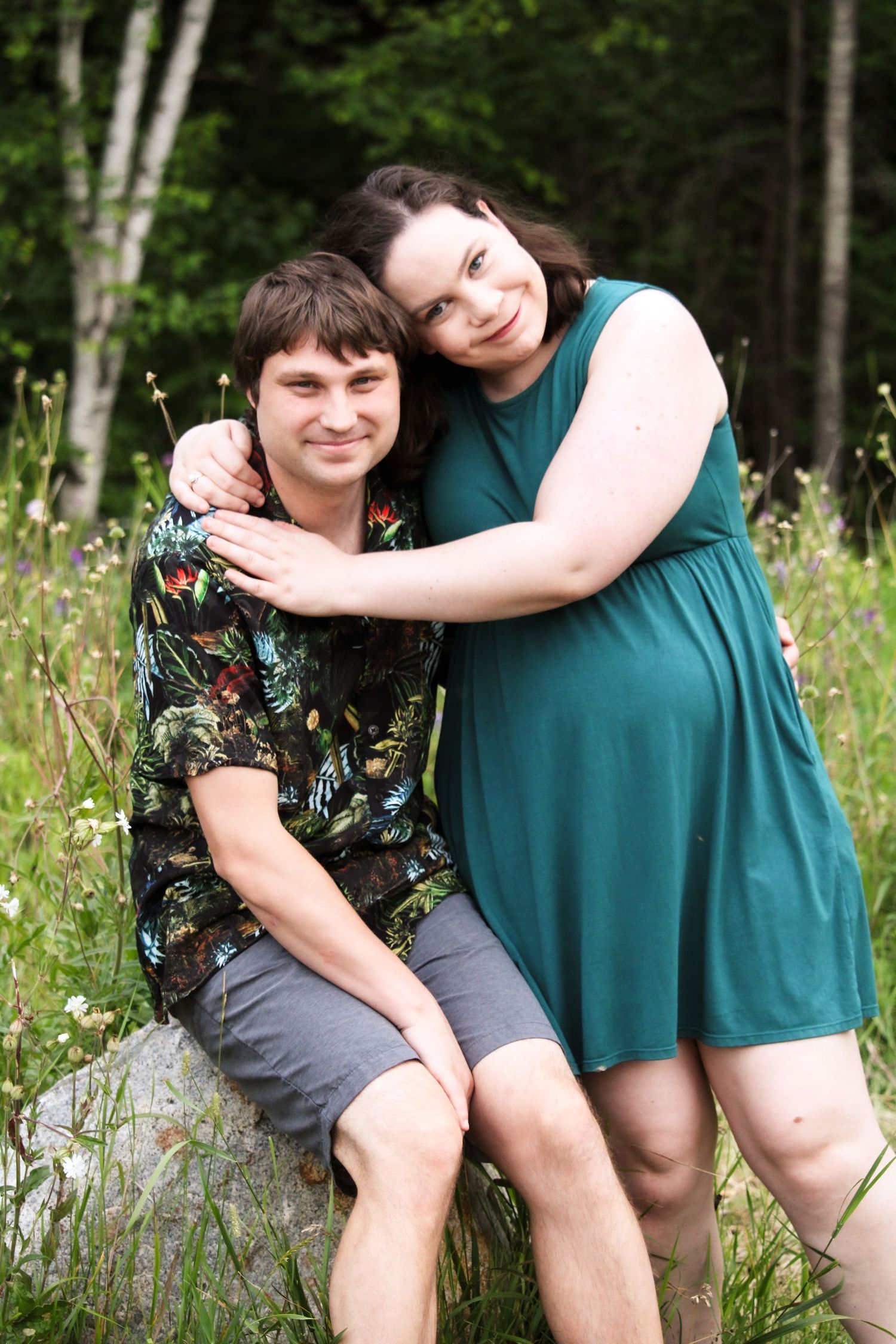 Woman in teal dress embraces man sitting on rock in a lush green meadow with wildflowers.