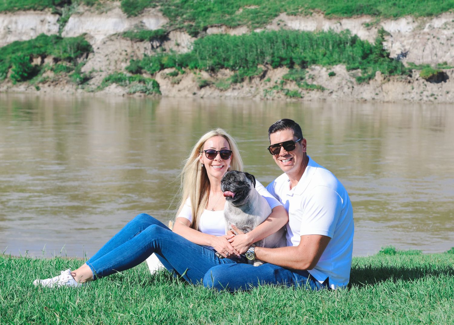Smiling couple sitting on grass by a river, holding a pug dog on a sunny day.