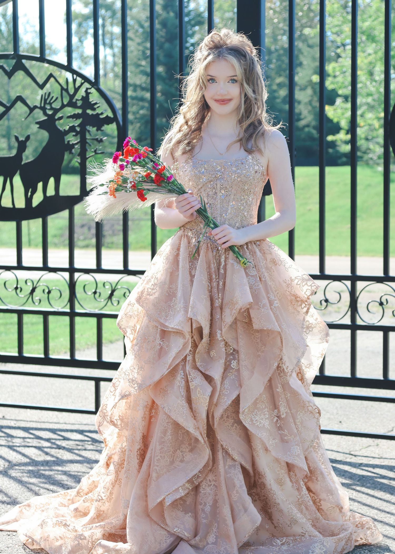 Young woman in elegant champagne gold ruffled ball gown holding colorful bouquet, standing before ornate black iron gates.