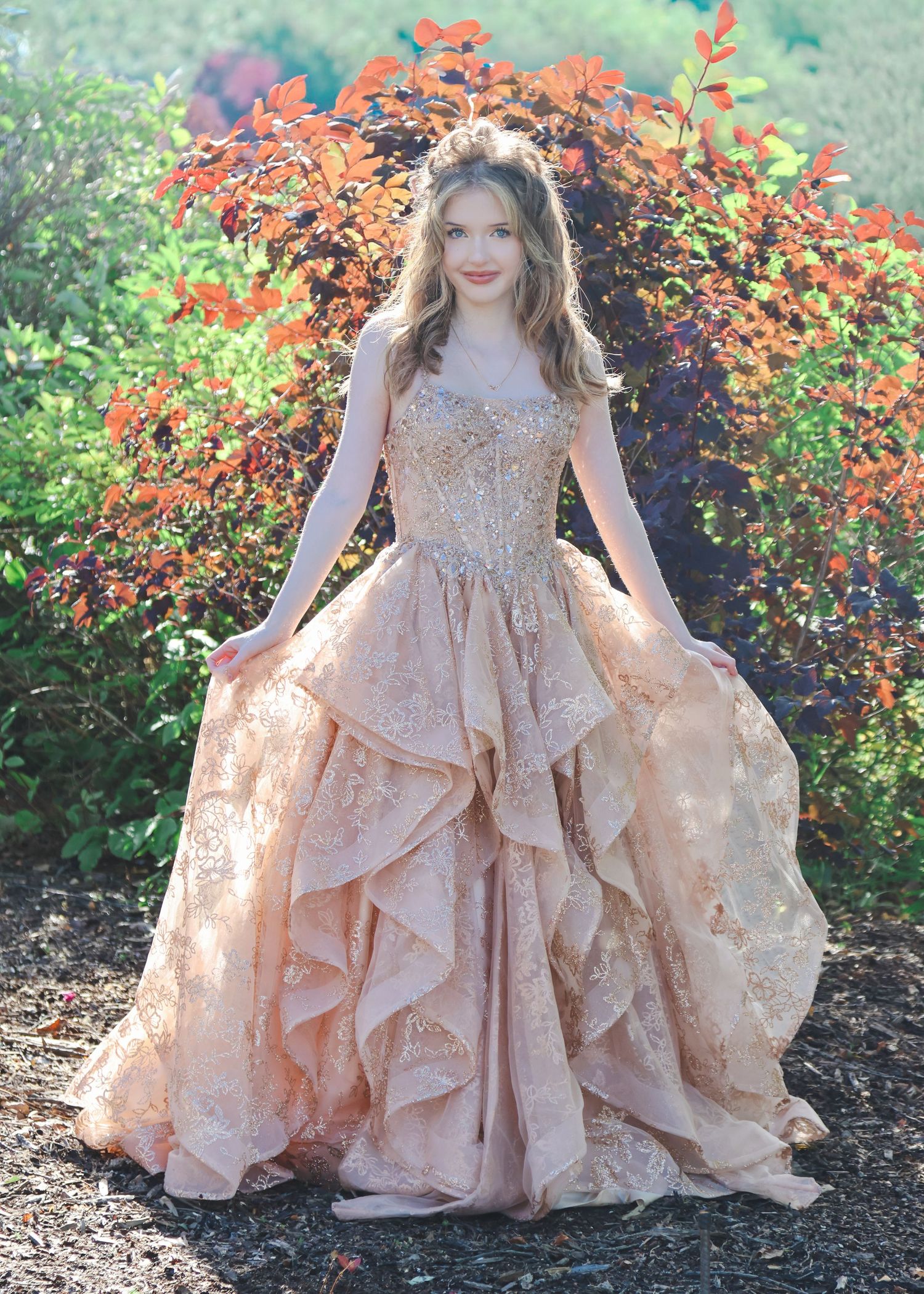 Young woman in elegant rose gold ruffled lace ballgown posing outdoors among vibrant autumn foliage.