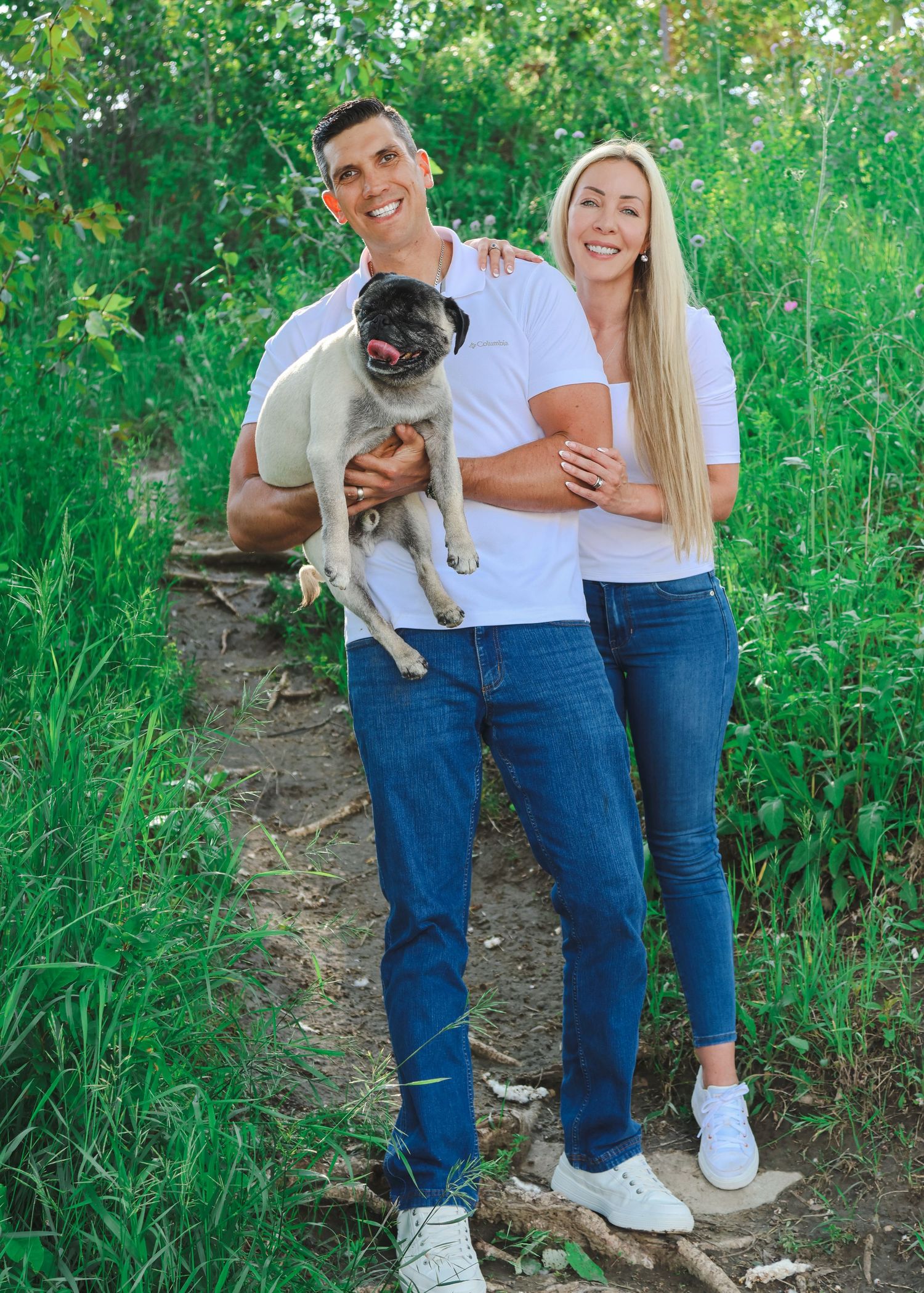 Smiling couple in white shirts and jeans holding a pug dog on a nature trail surrounded by green foliage.