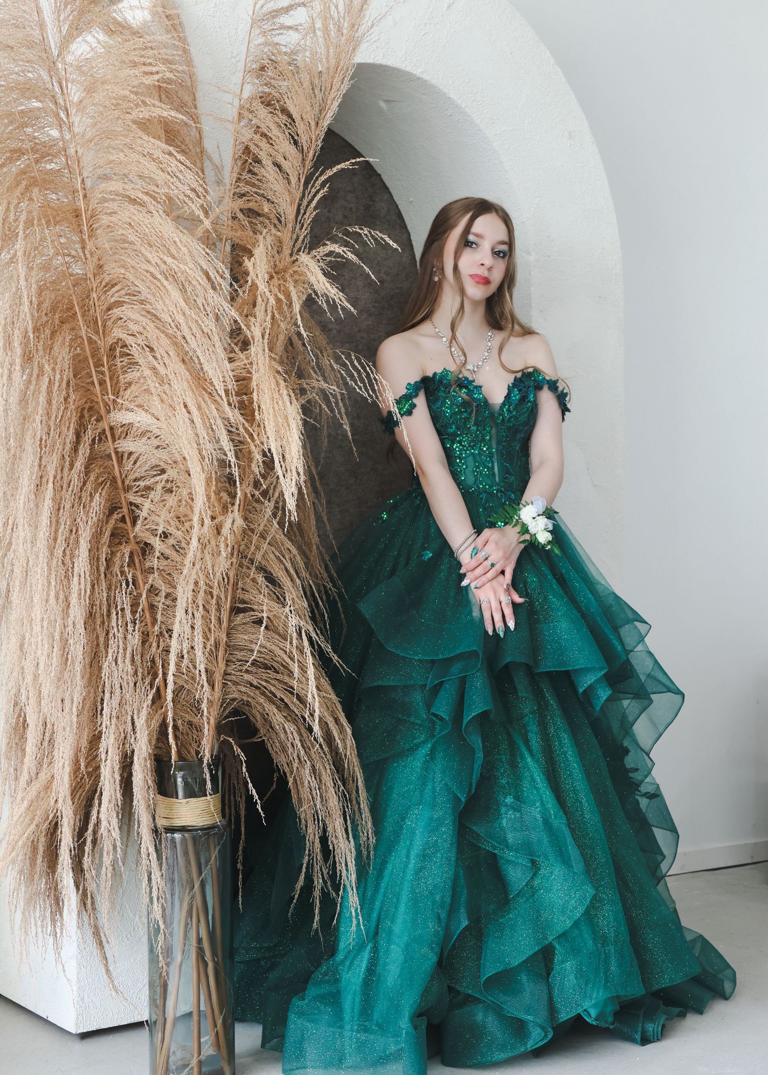 Woman in stunning emerald green off-shoulder ball gown with floral details posing near pampas grass.