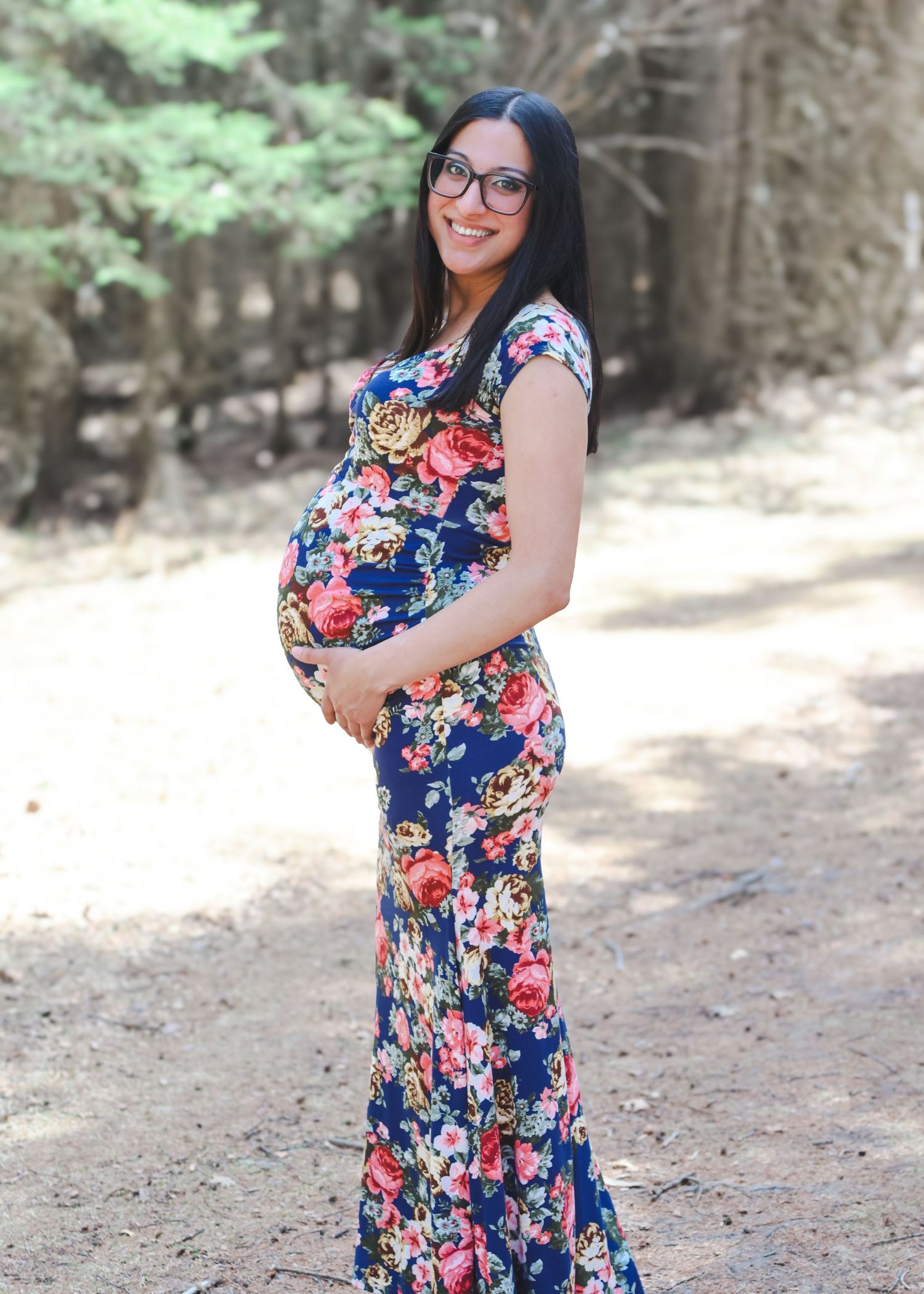 Smiling pregnant woman in navy floral maxi dress holding baby bump on sunny outdoor forest path.