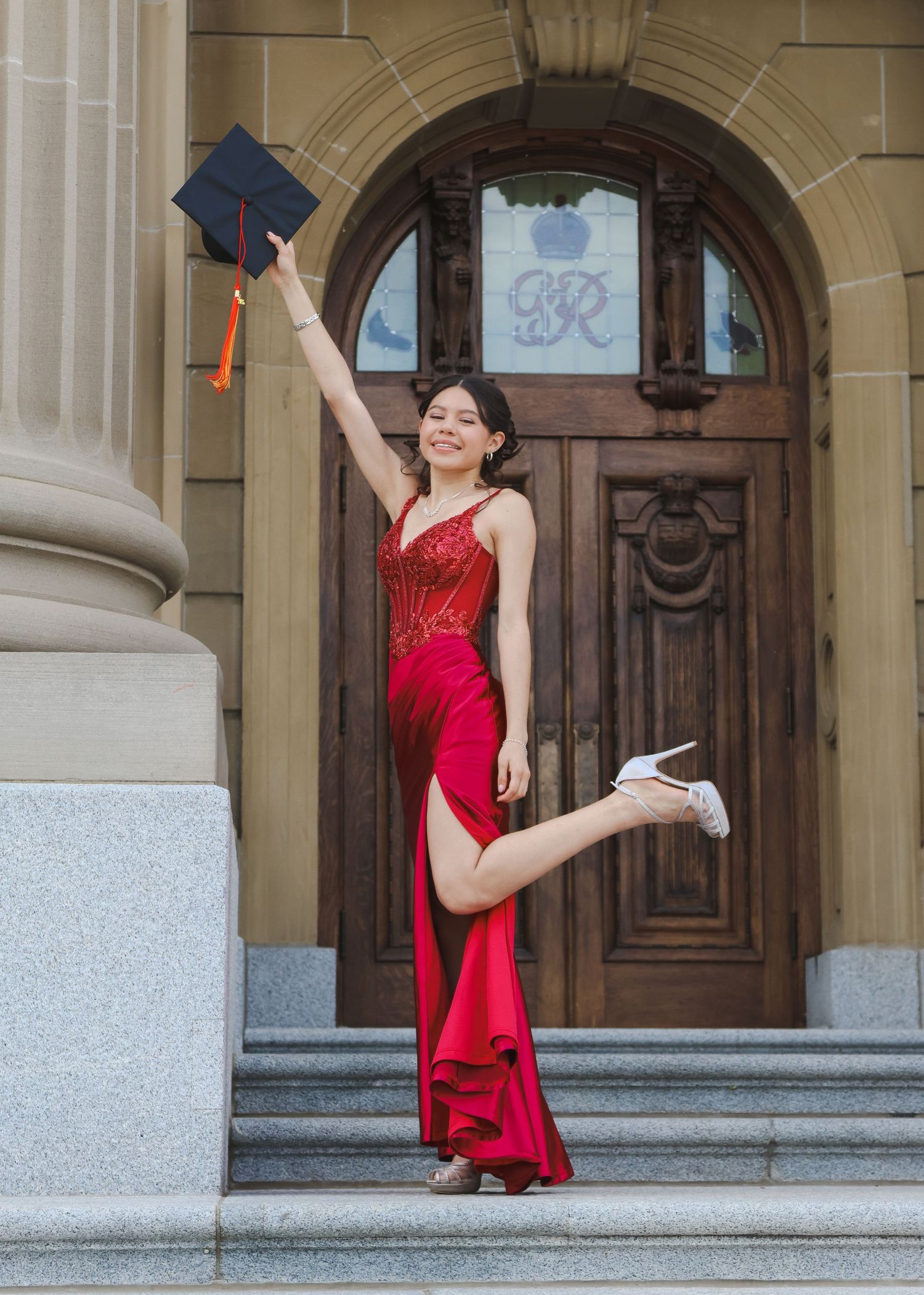 Joyful graduate in red gown tossing cap outside ornate wooden doors.