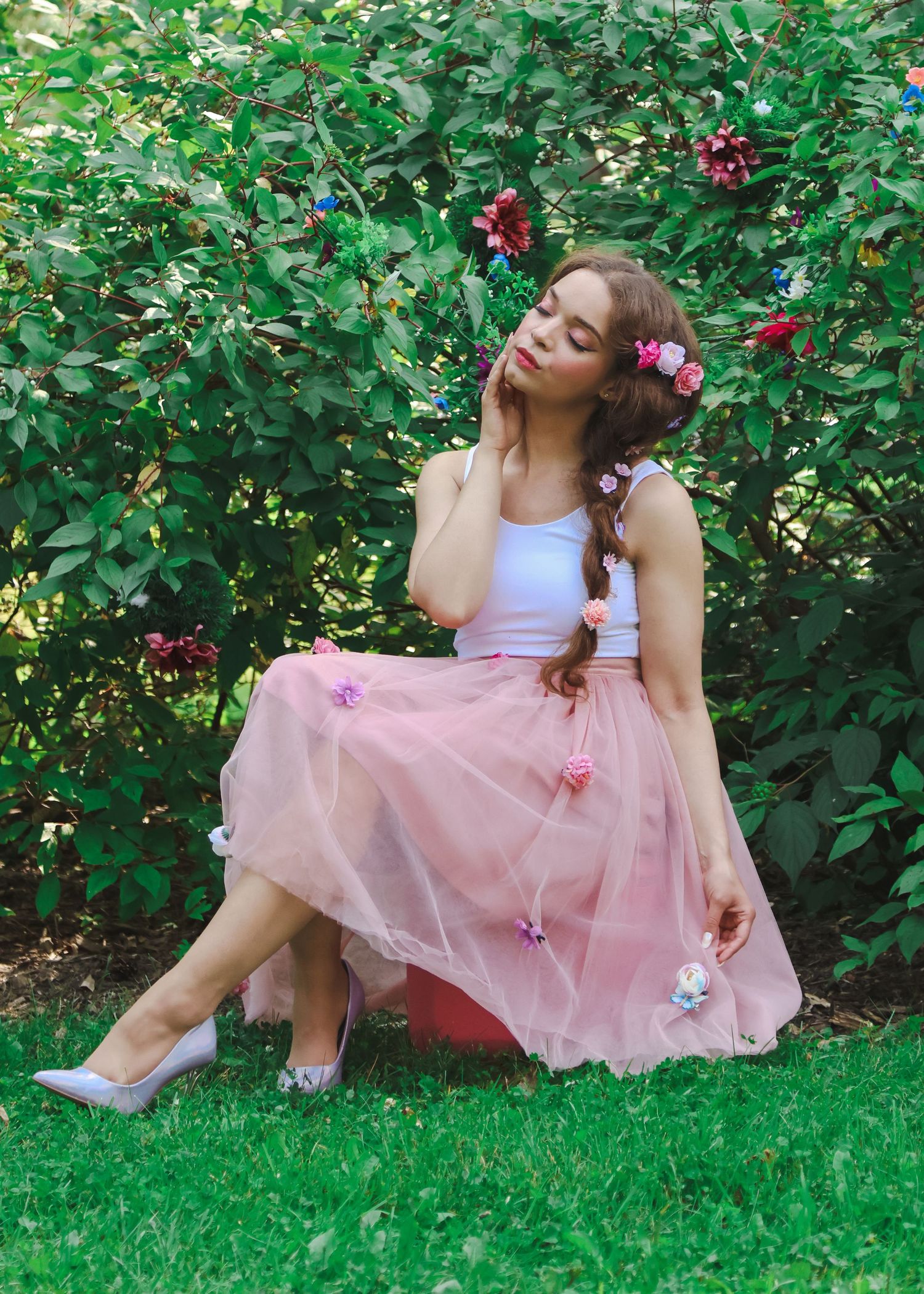 Young woman in pink tulle skirt with floral hair accessories sits gracefully on grass near lush green bushes.