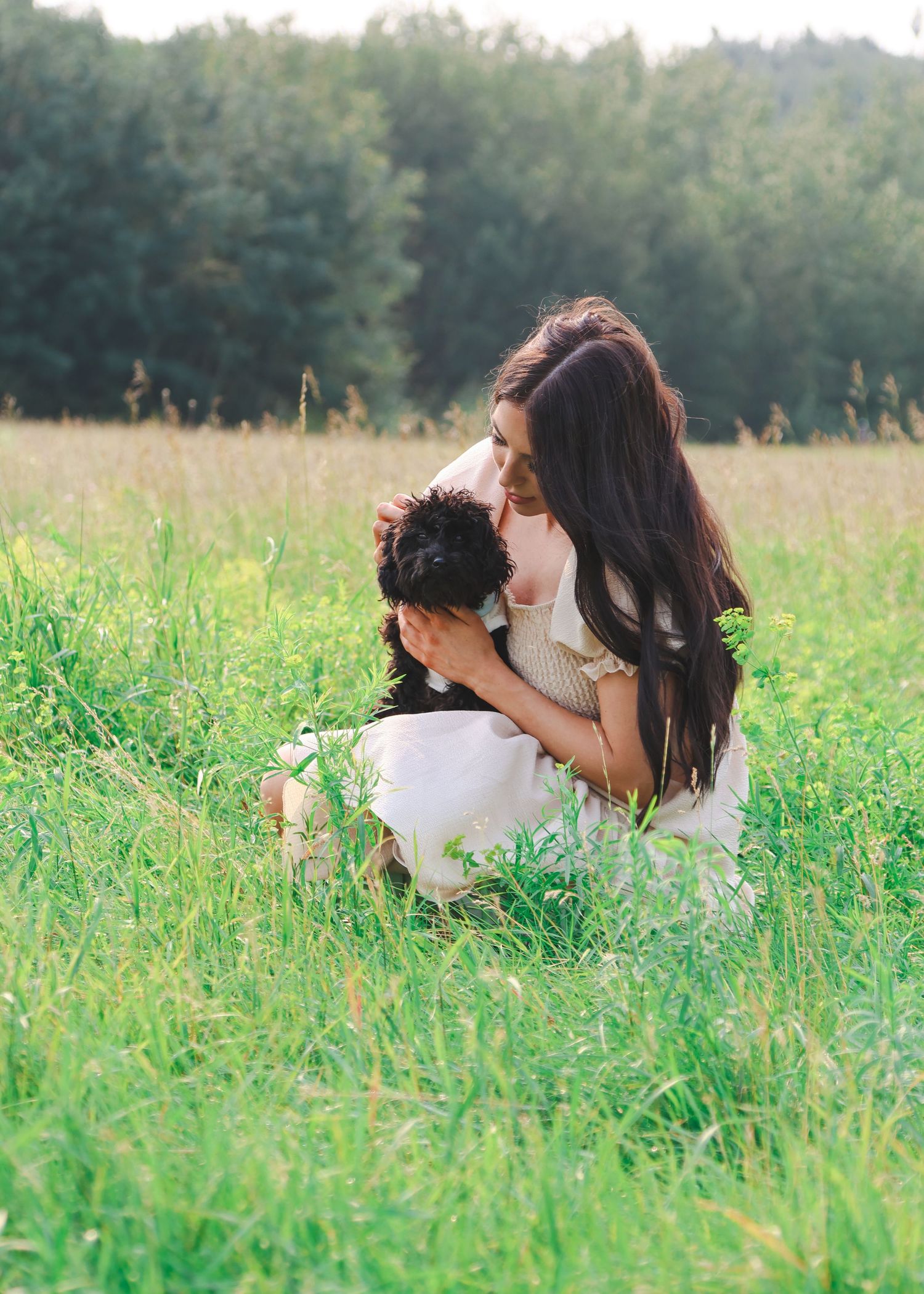 Young woman with long dark hair crouching in green field, tenderly holding a small black puppy.