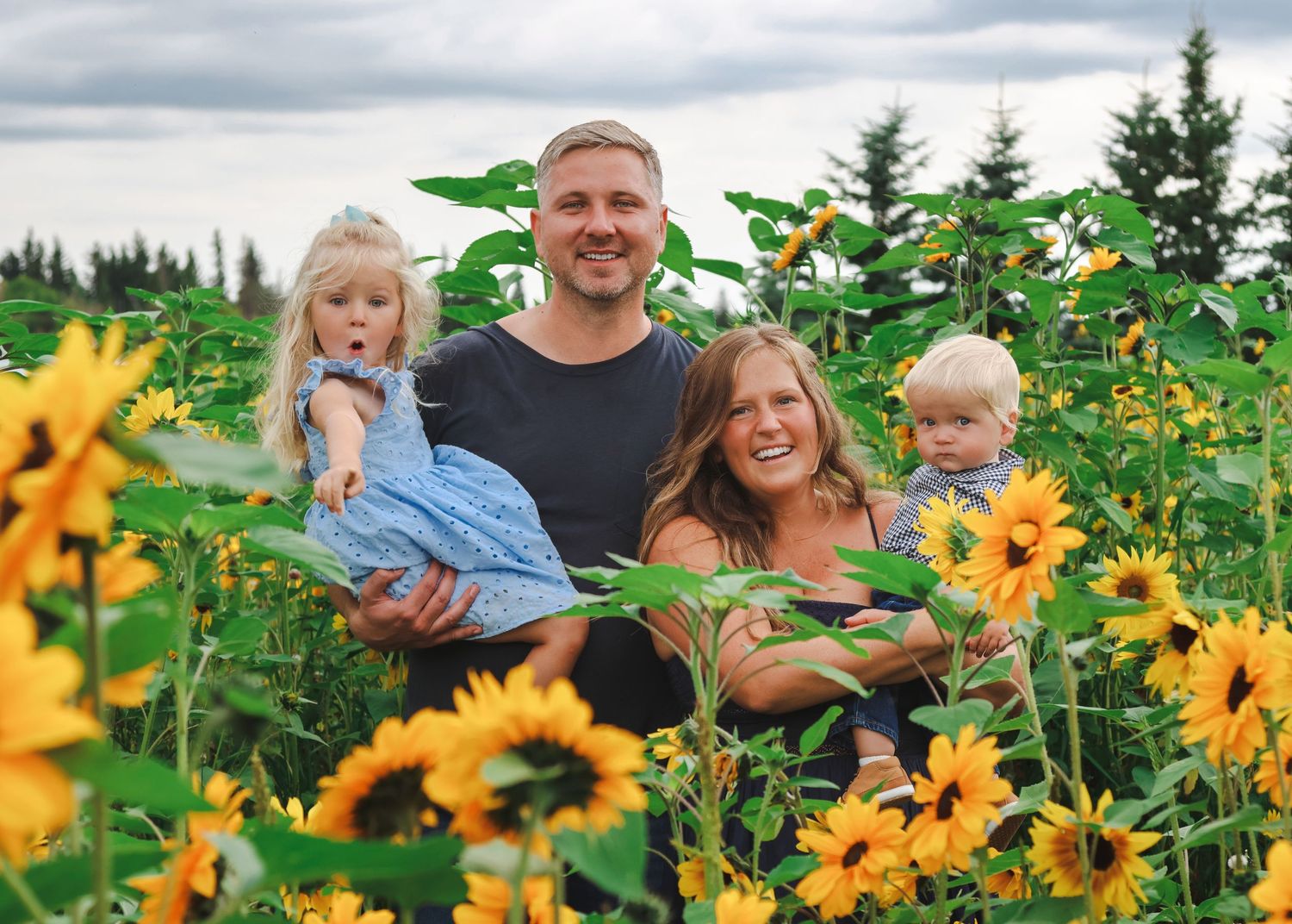 Family of four smiling in a vibrant sunflower field on a cloudy day, parents holding two young children.