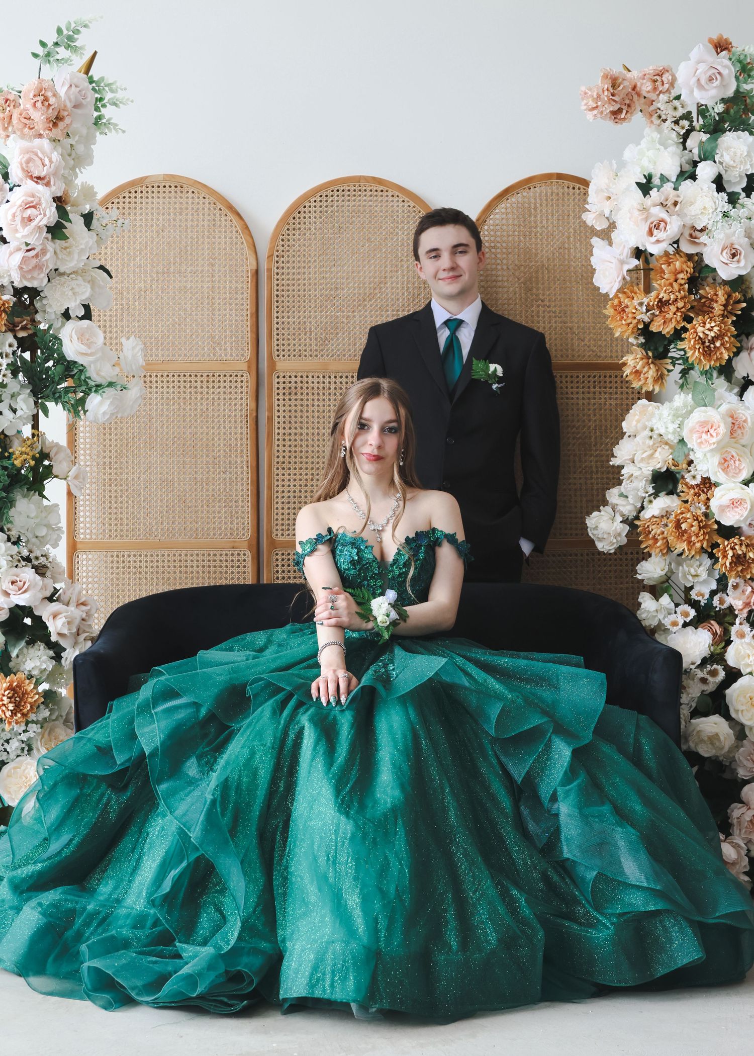 Couple posing with woman in emerald green ball gown seated before rattan backdrop with flowers.