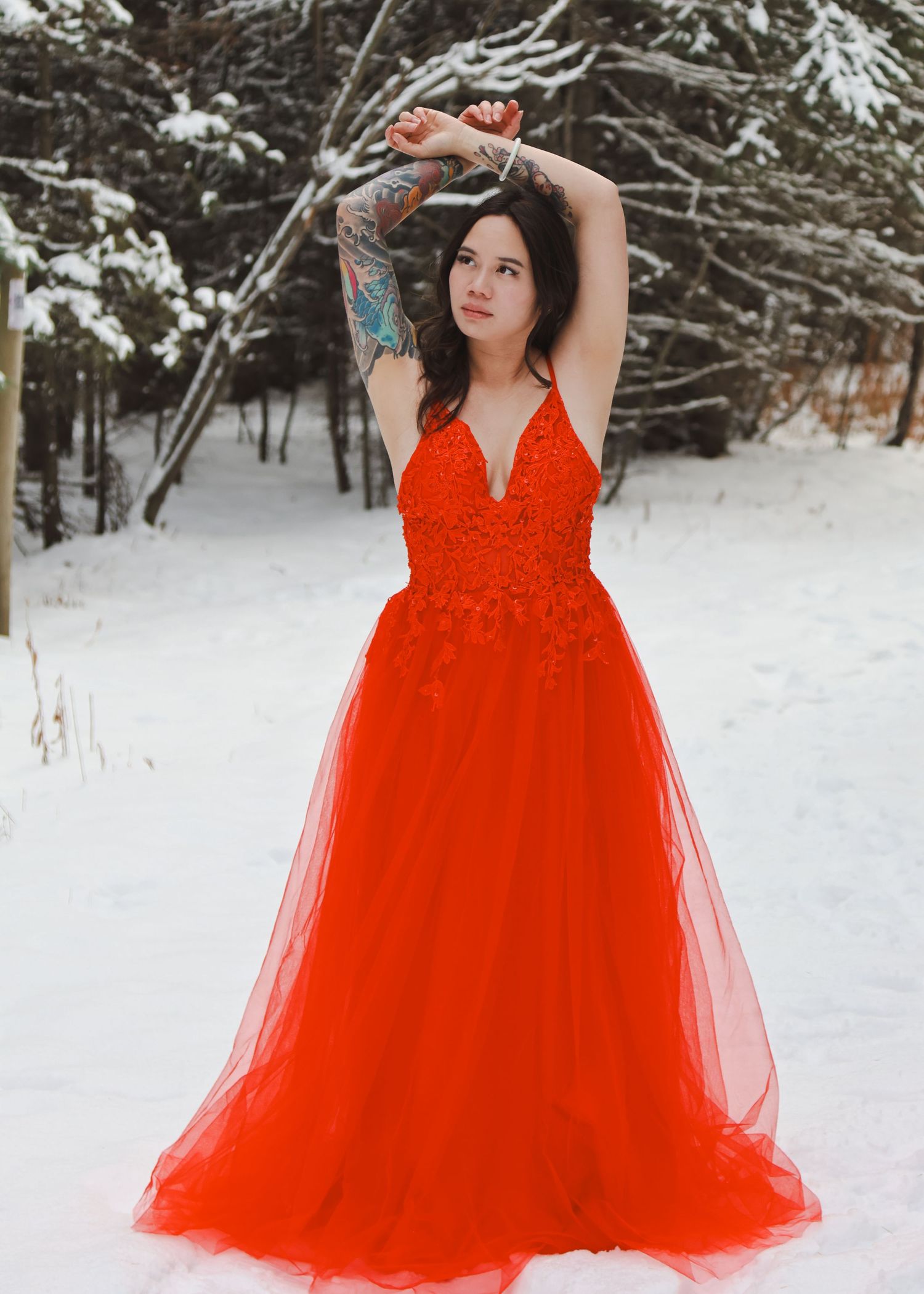 Woman in red lace ballgown poses gracefully in snowy winter forest, showcasing tattoos and elegant formal dress.