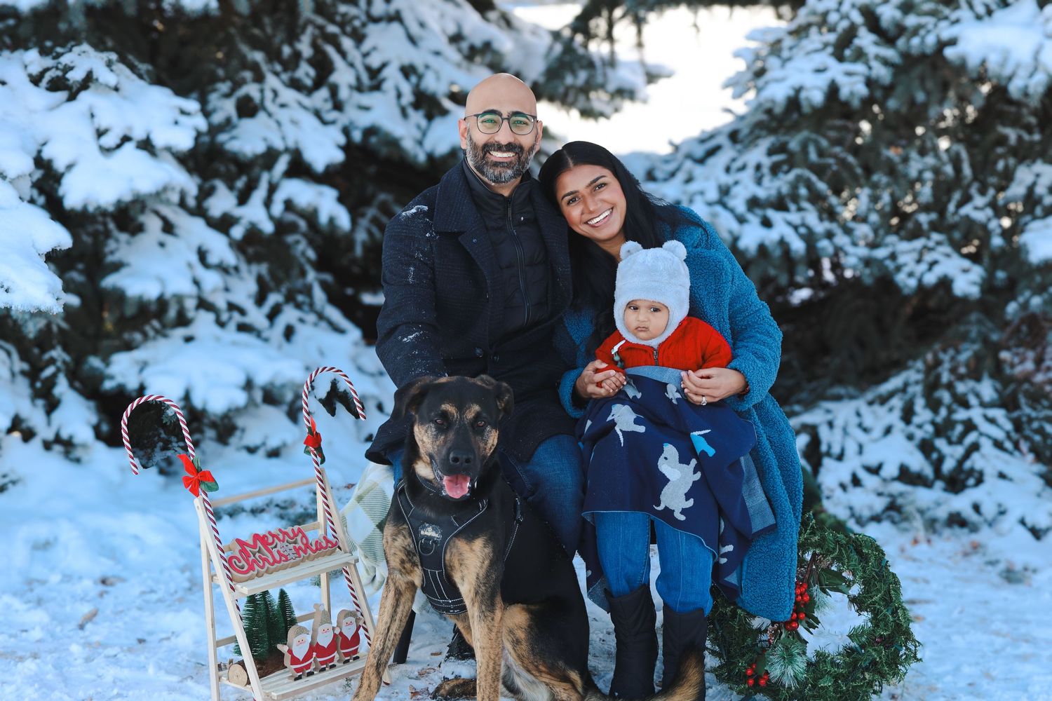 Family portrait with dog in snowy winter wonderland with festive sled and wreath decorations.