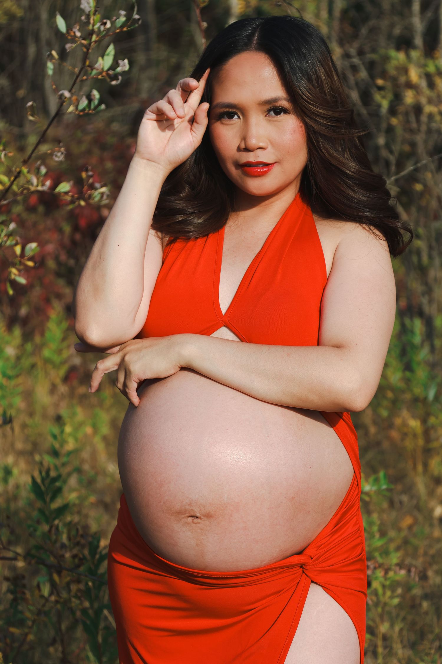 Pregnant woman in red halter dress cradling bare baby bump outdoors in nature.