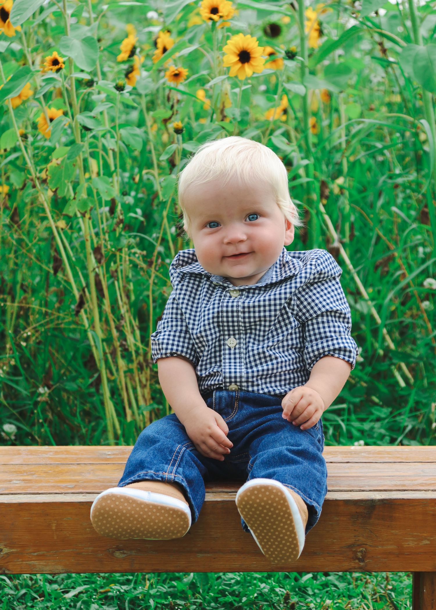 Smiling blonde toddler boy in plaid shirt and jeans sitting on wooden bench with sunflowers in background.