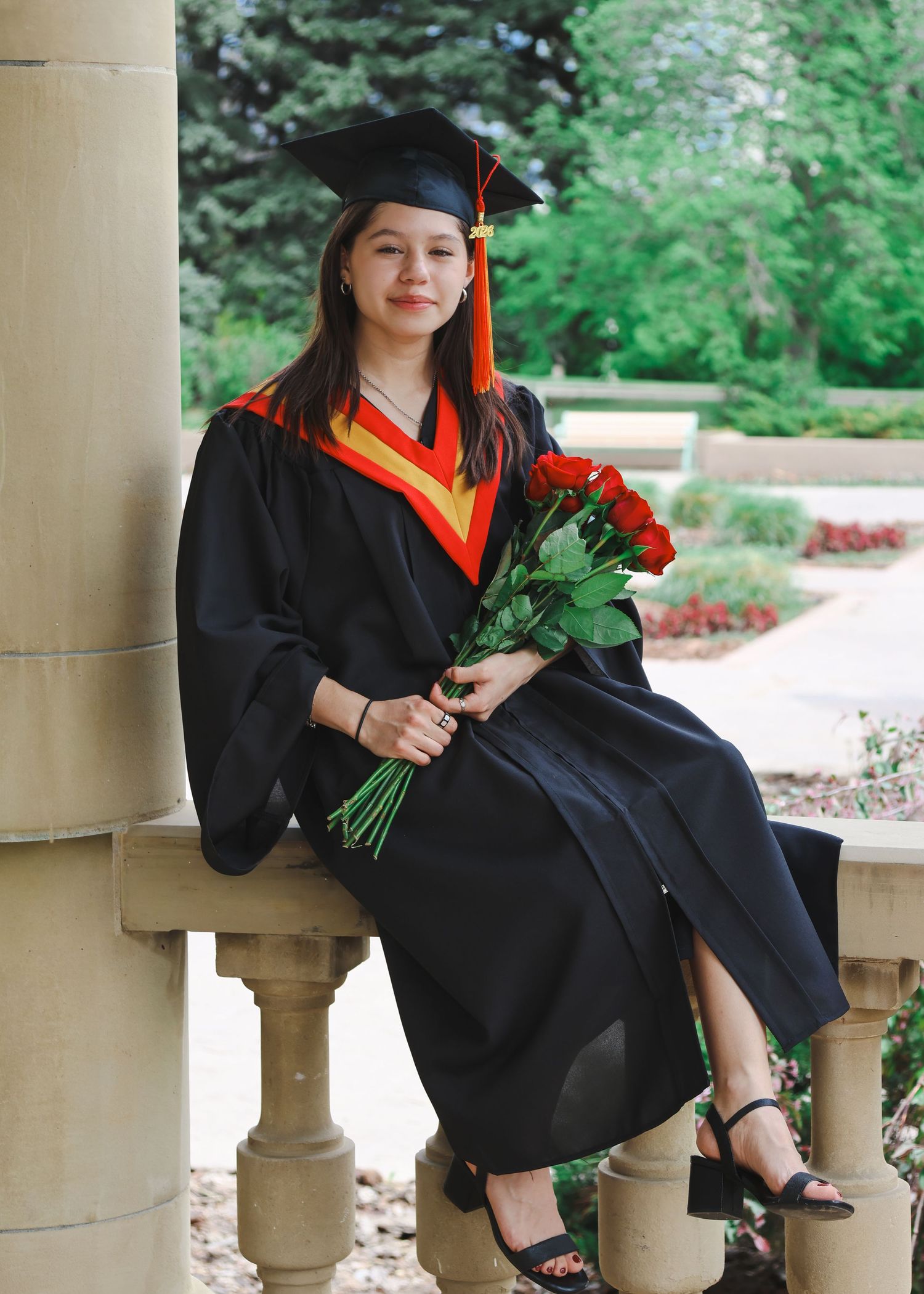Smiling graduate in black cap and gown holding red roses seated on balustrade.