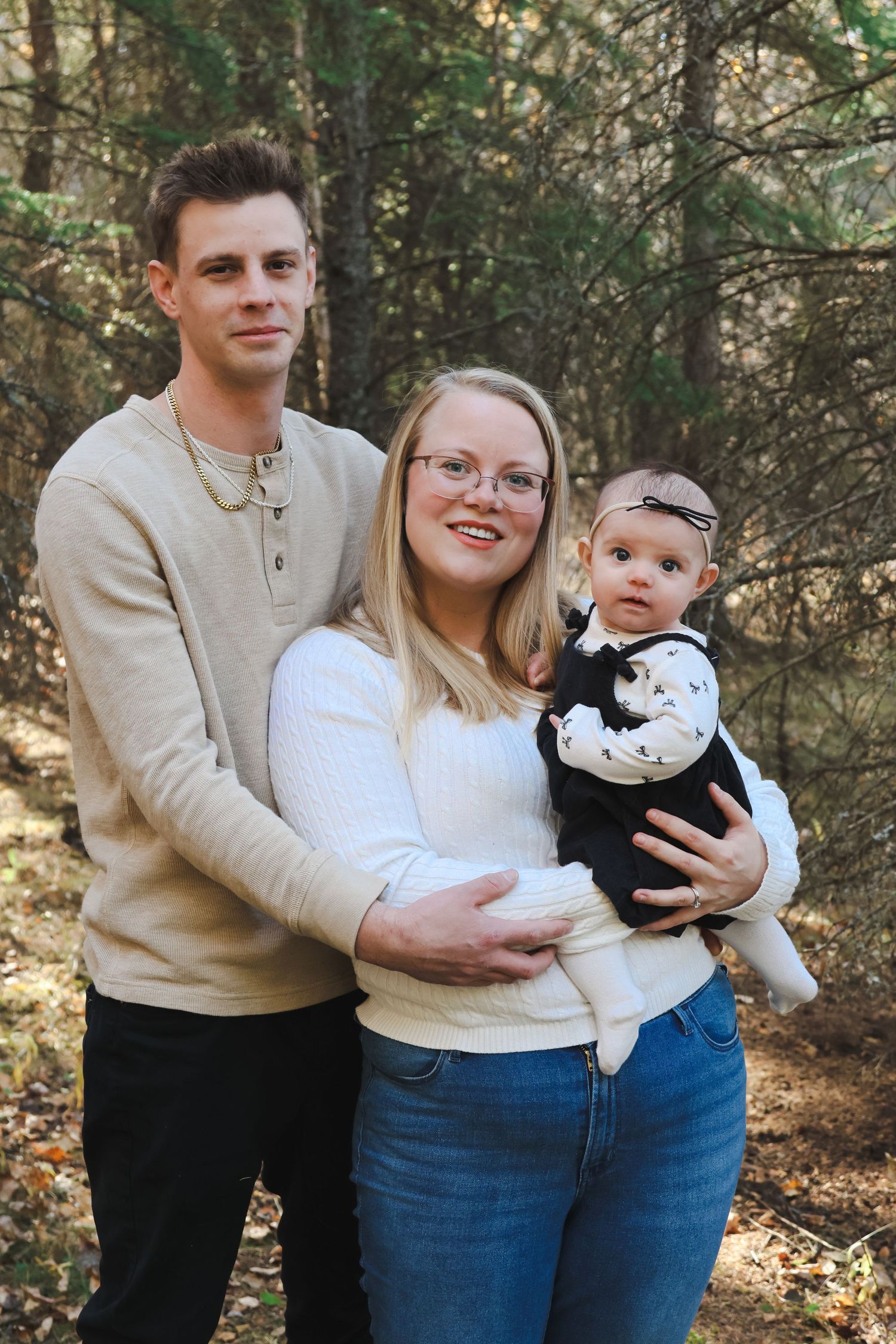 Young family with baby poses in autumn forest, parents in neutral tones holding infant in black outfit.