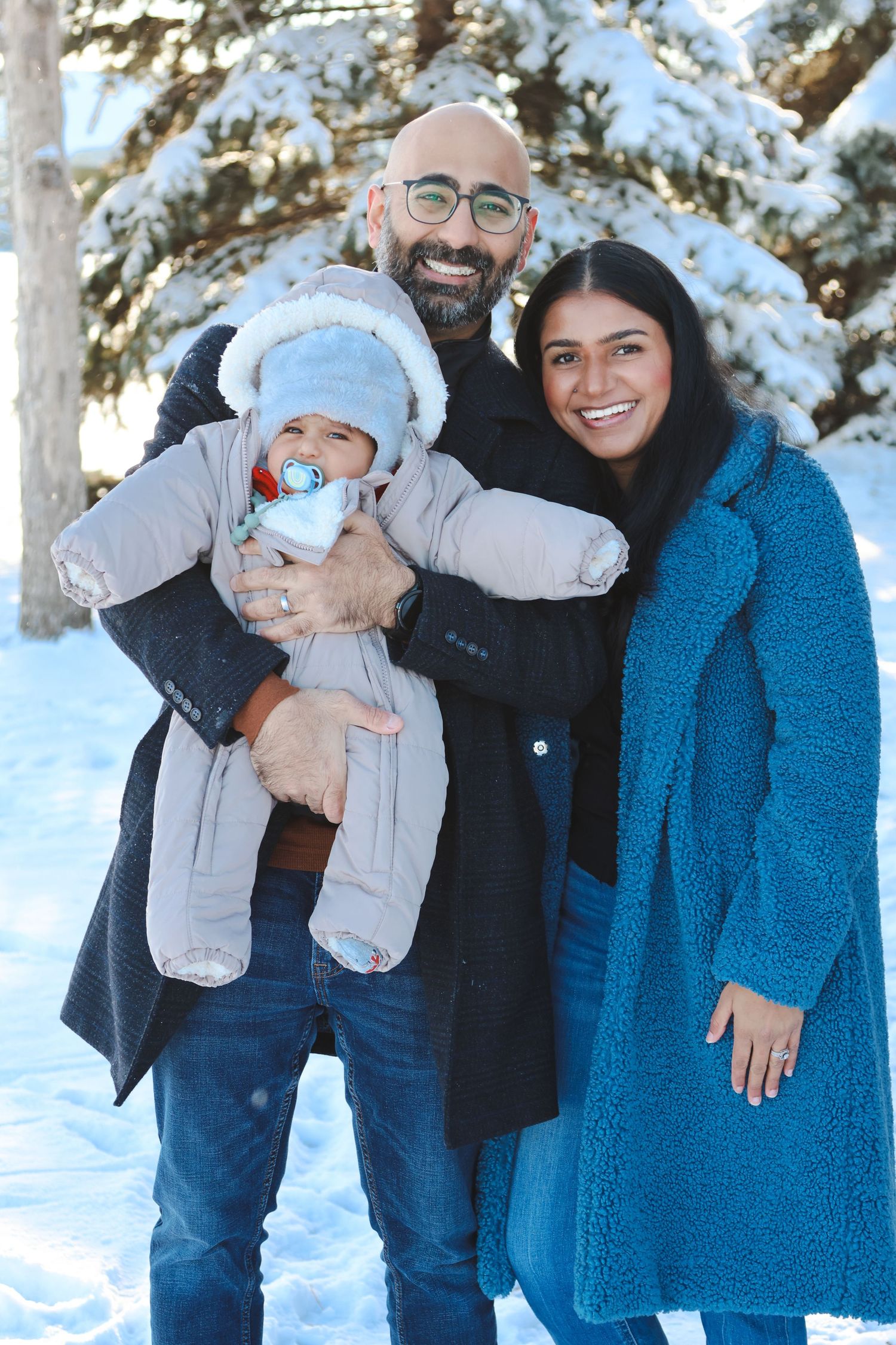 Smiling couple holds bundled baby outdoors in snowy winter park setting.