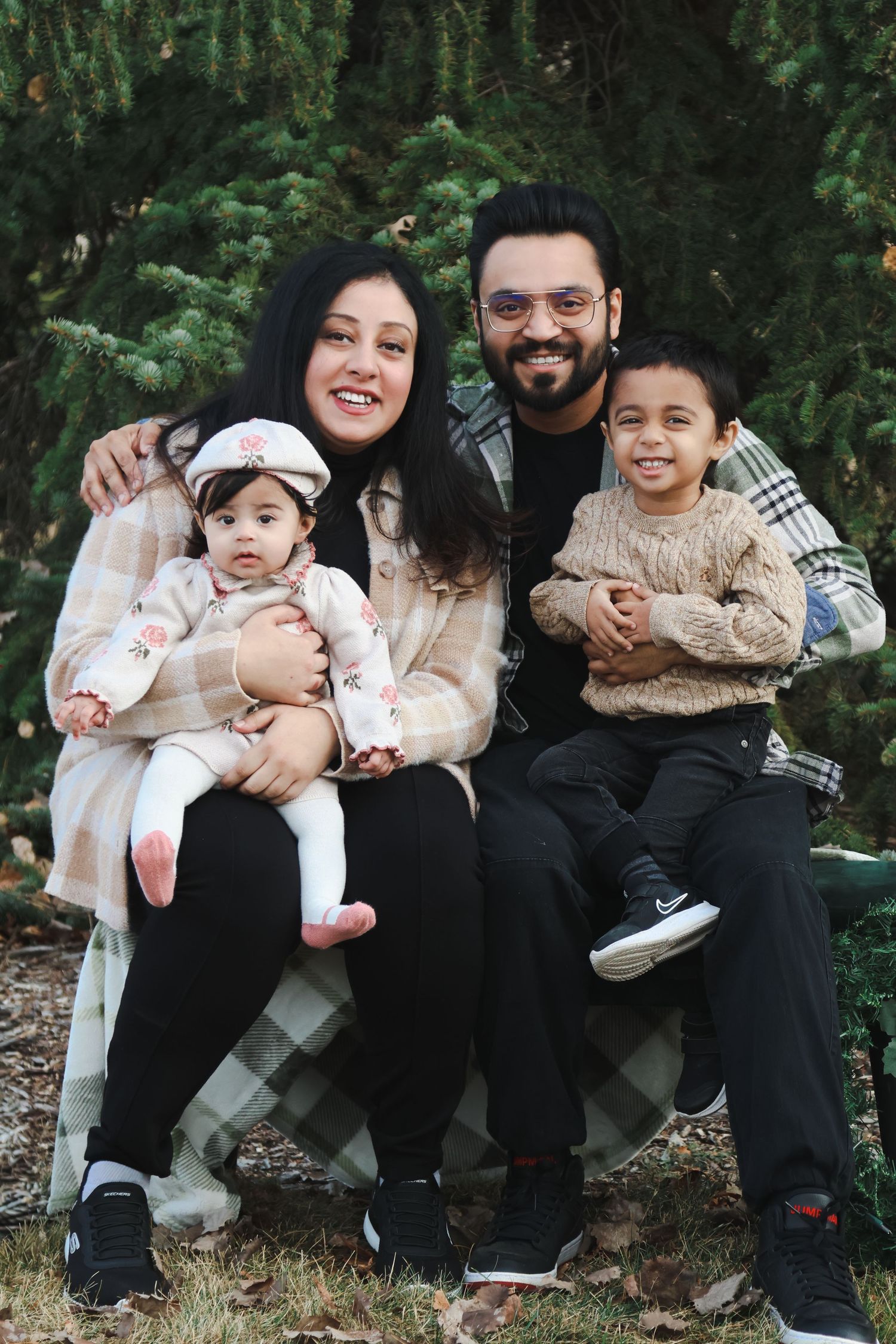 Smiling family of four sits together near evergreen trees, parents holding young baby and toddler.