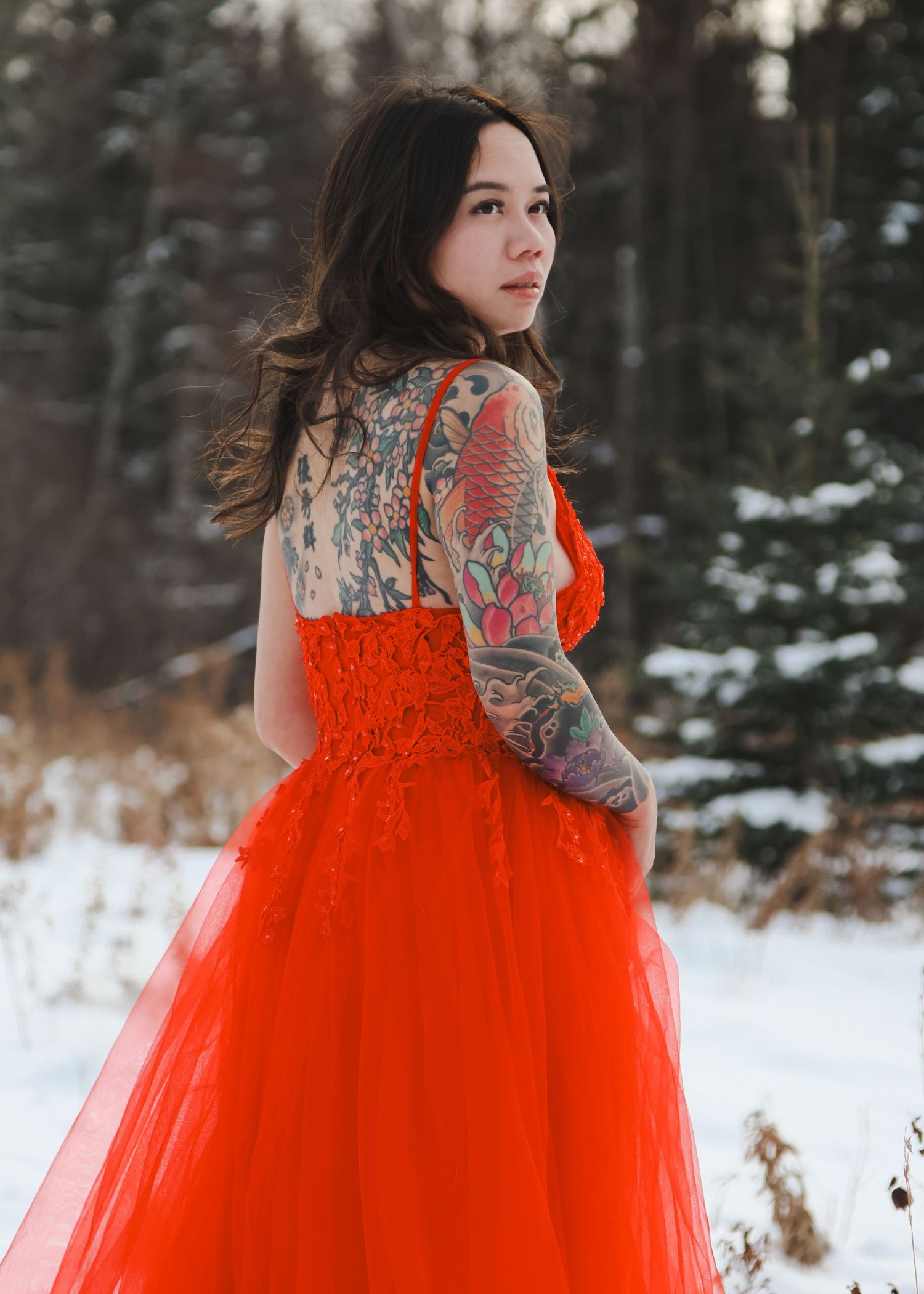 Woman in red lace gown with colorful tattoos posing by birch tree in snowy winter setting.