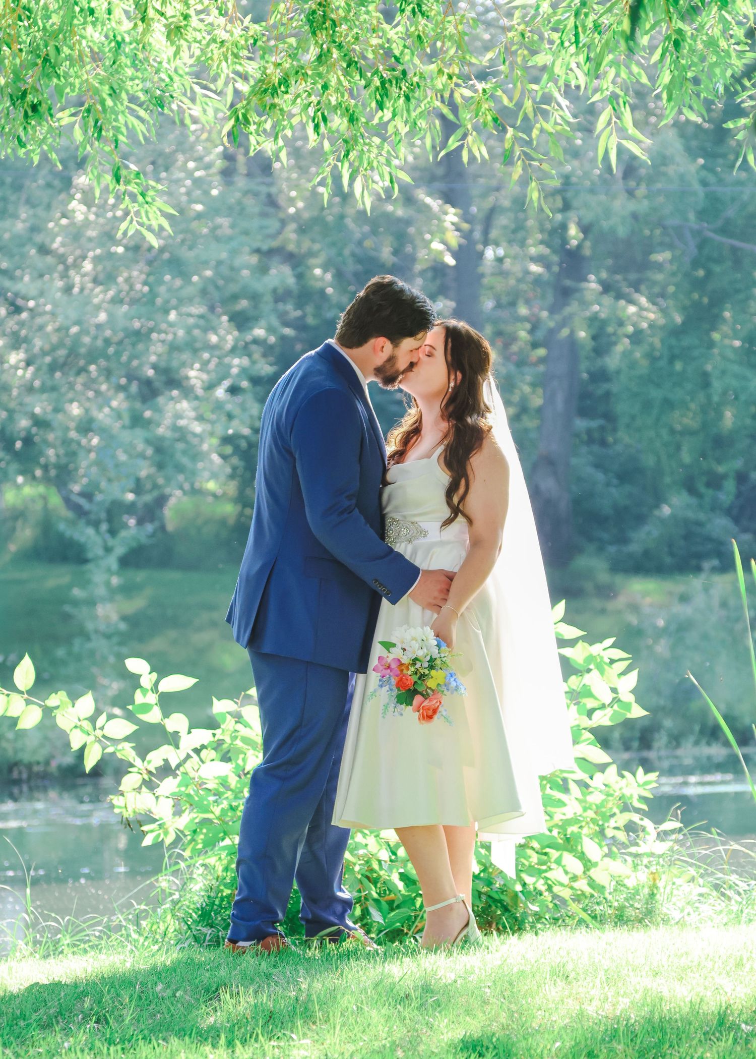 Bride and groom nearly kissing under weeping willow tree beside tranquil water.