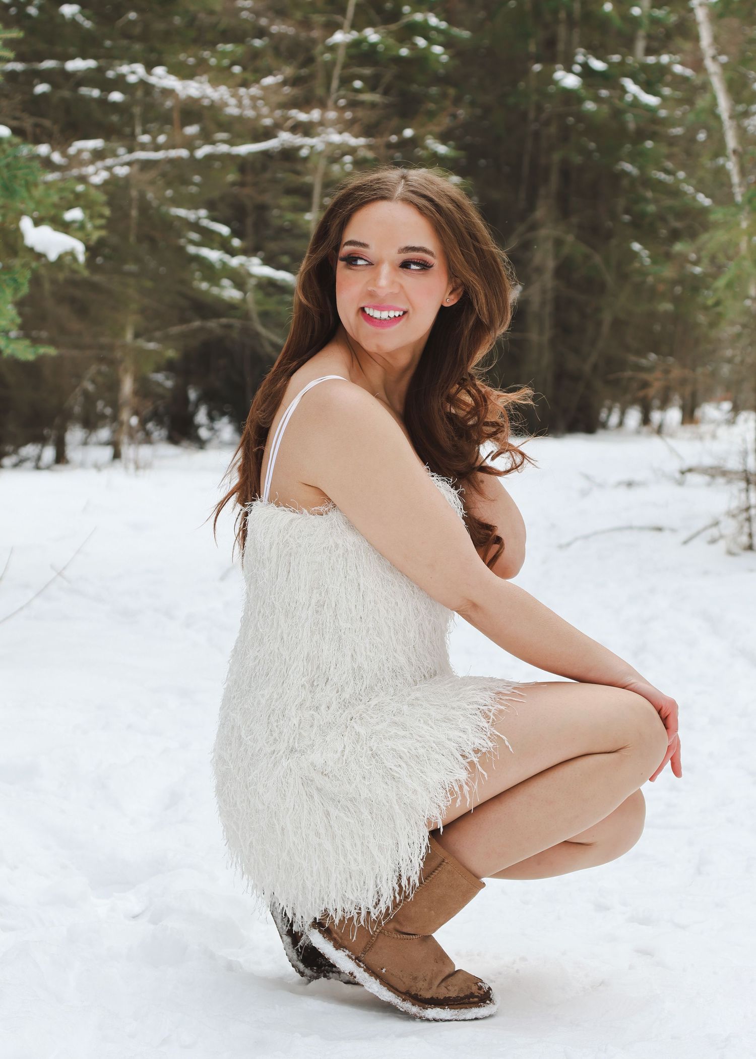 Young woman in white fur dress and brown boots crouching in snowy forest, smiling at camera.