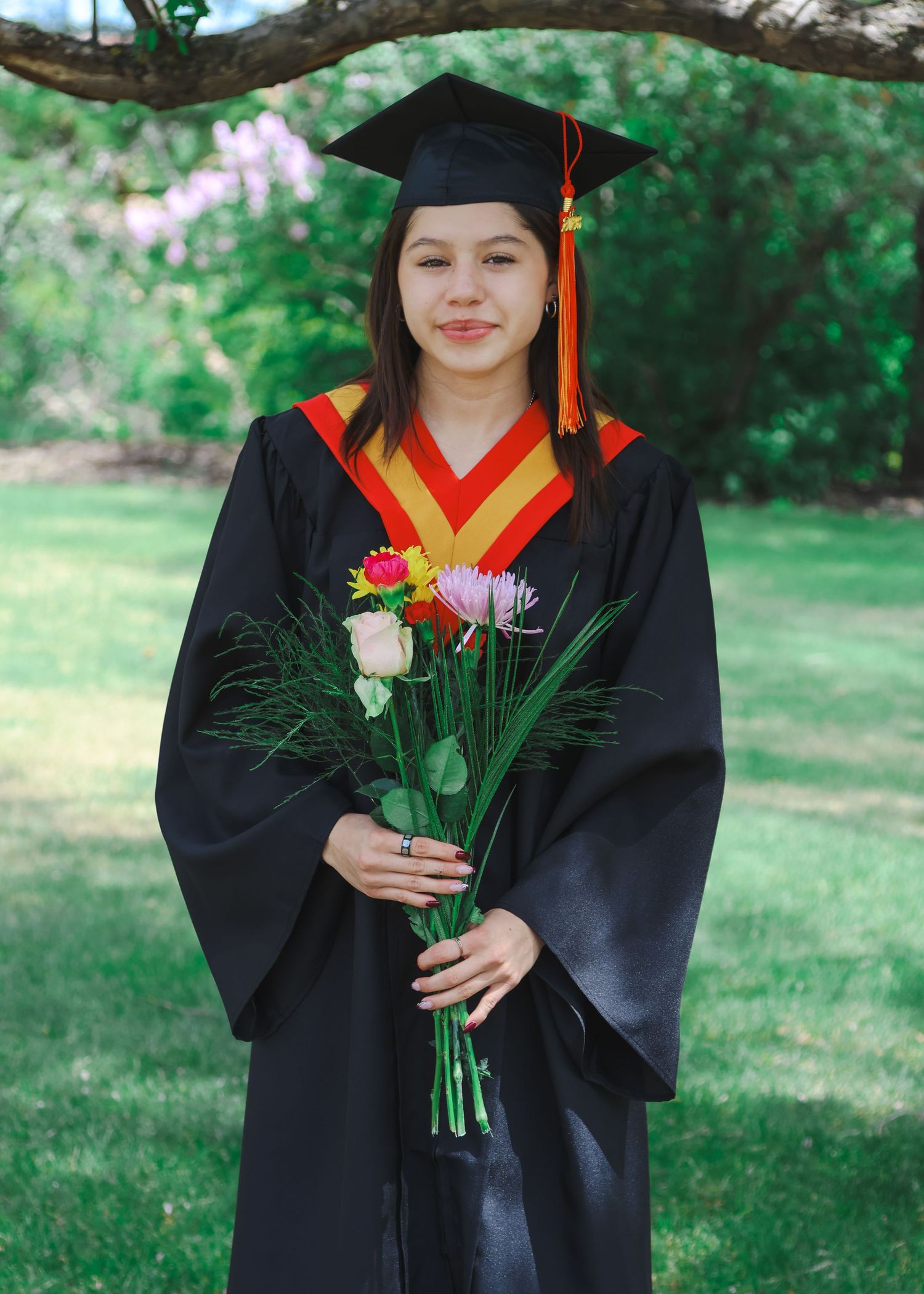 Graduate in black cap and gown holding colorful bouquet in lush green garden.