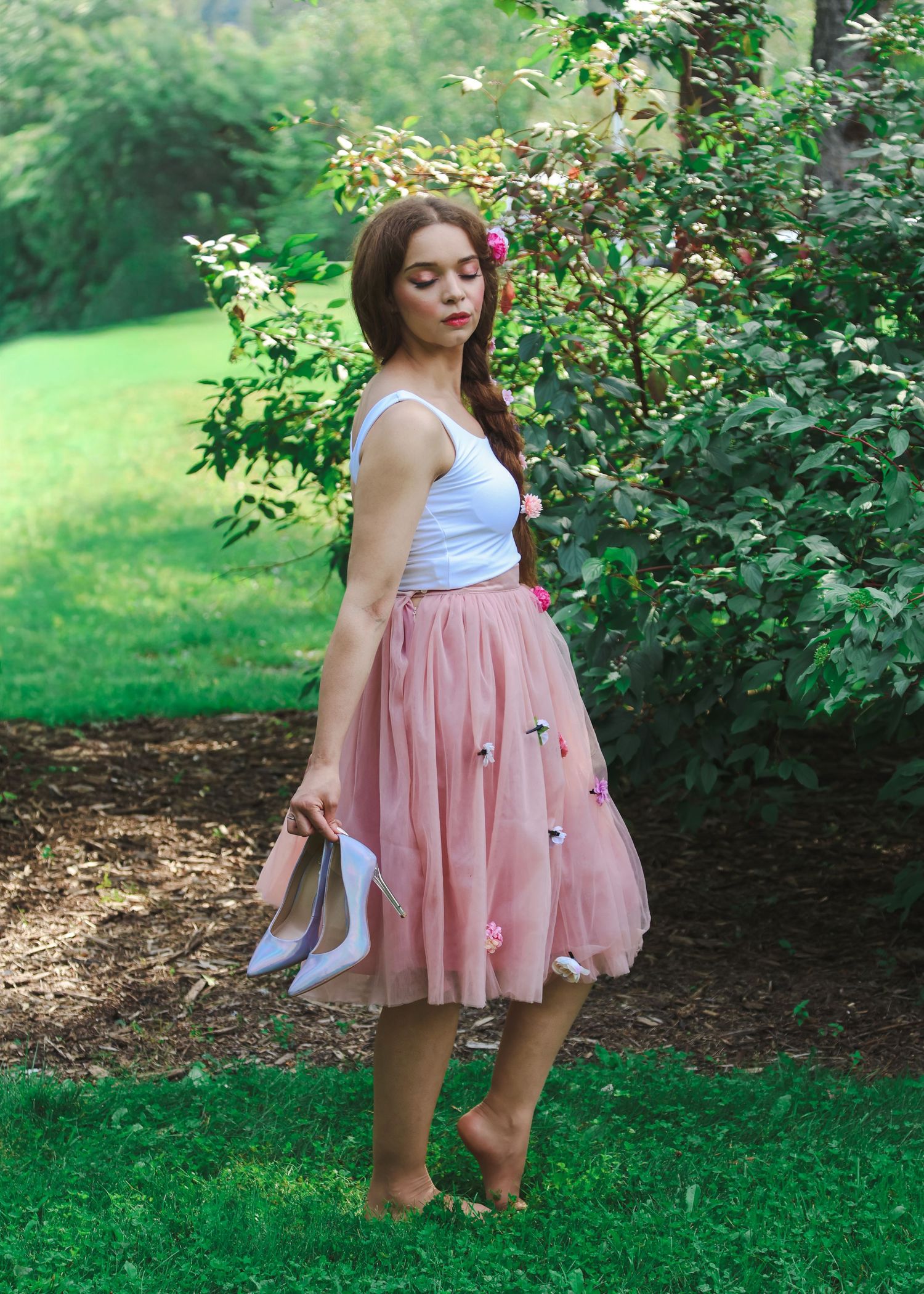 Young woman in white top and pink tulle skirt holds heels barefoot outdoors near lush green bushes.
