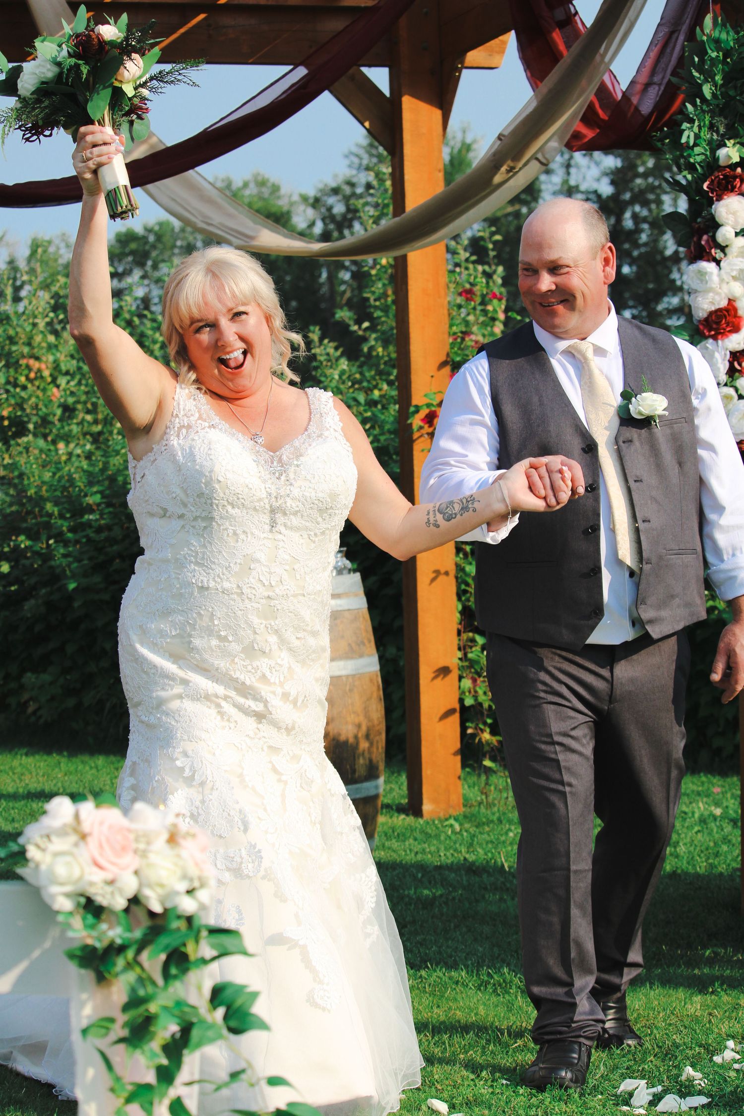 Joyful bride raises bouquet while groom smiles beside her at outdoor wedding ceremony under wooden arch.