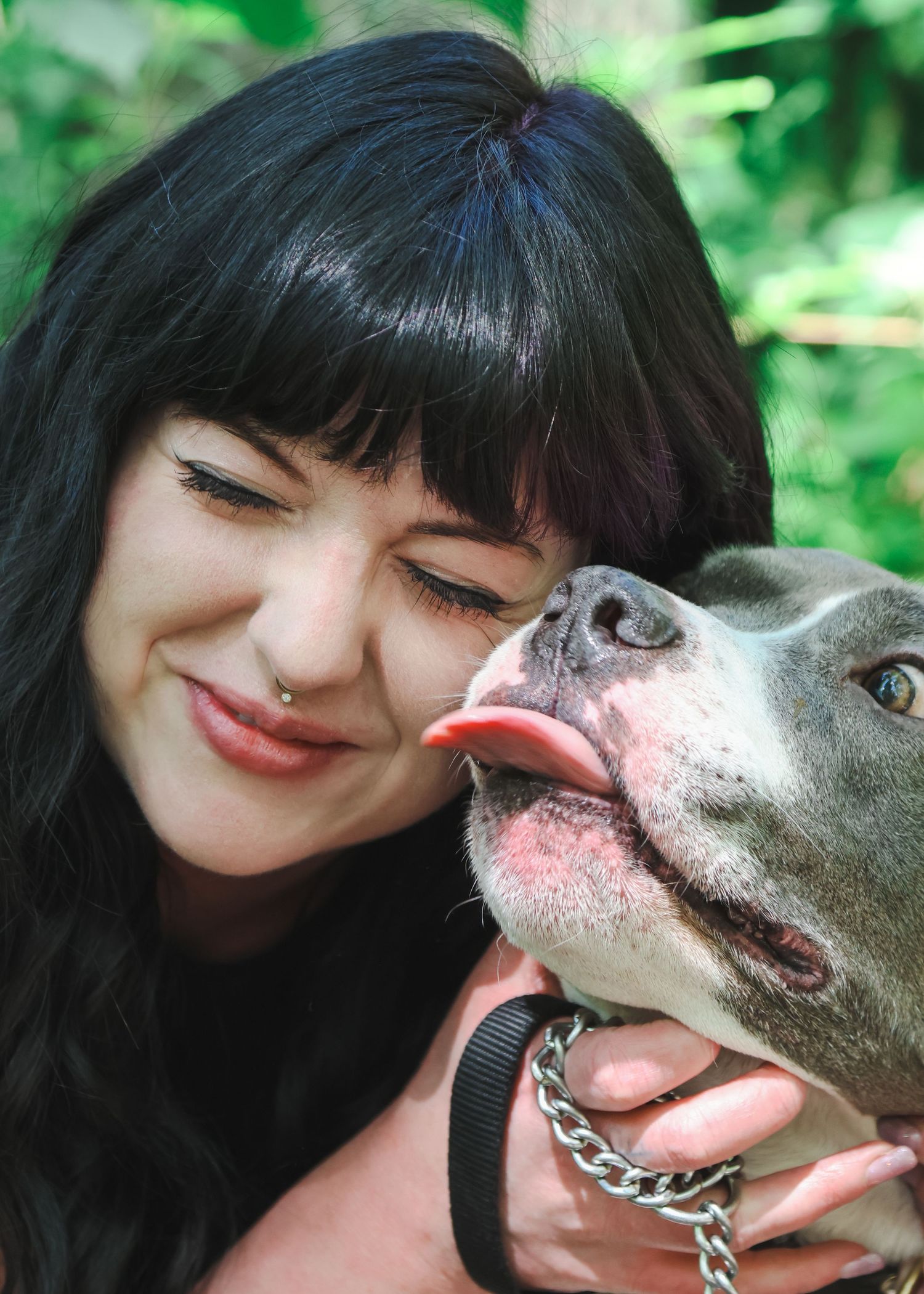 Woman with black bangs smiling as a gray pit bull dog licks her cheek outdoors.