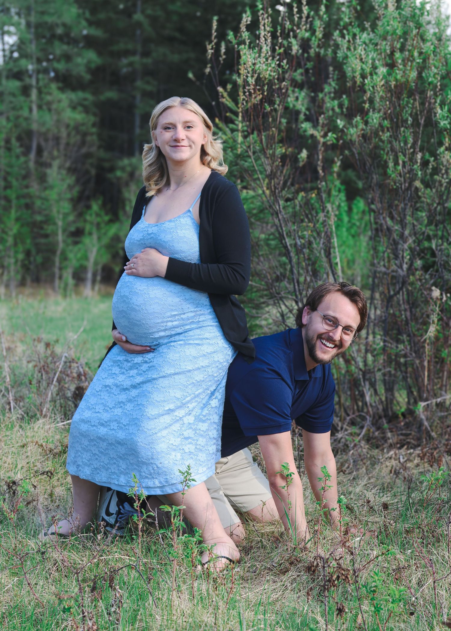 Laughing expecting couple posing playfully outdoors, man kneeling behind pregnant woman in blue dress.