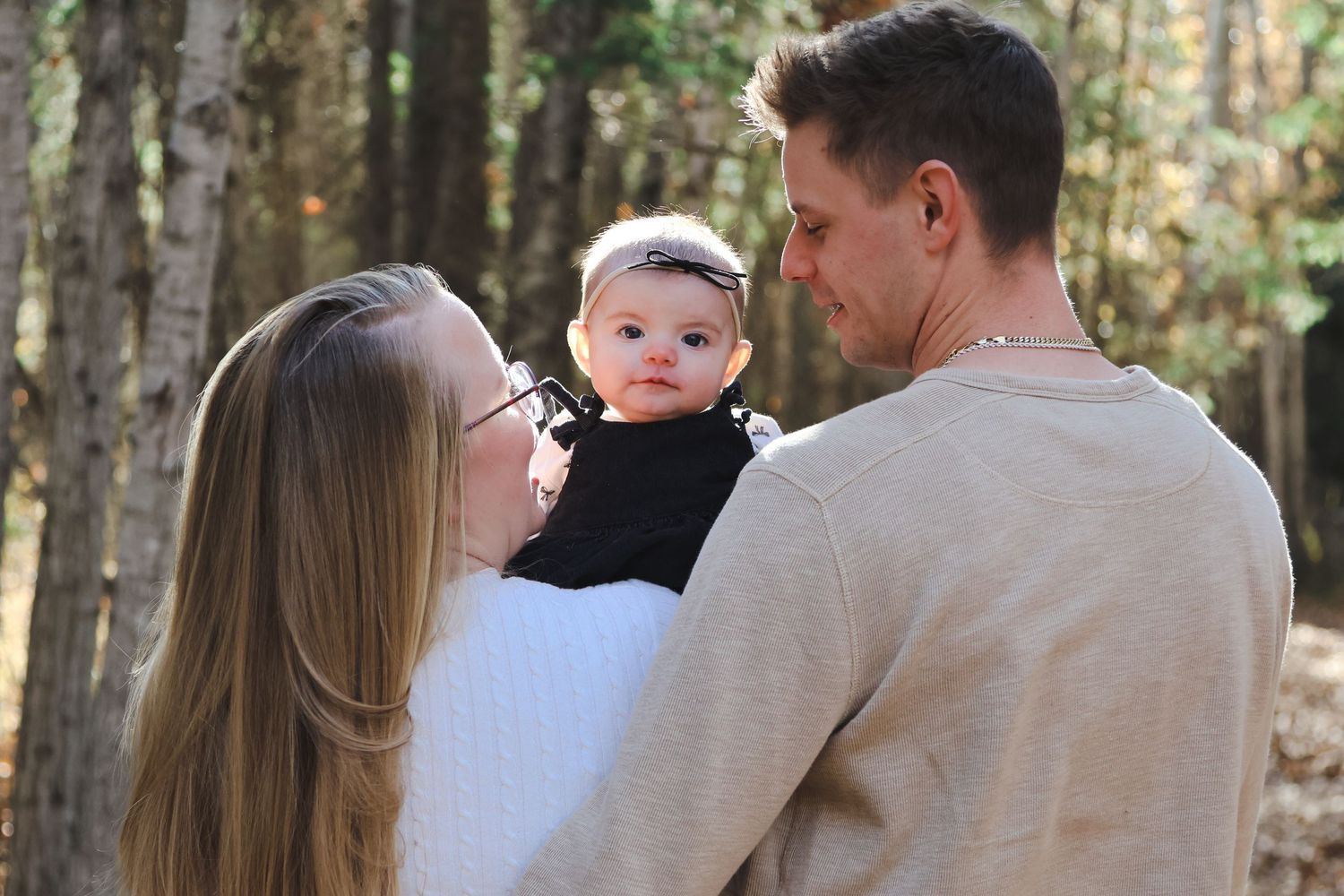 Young couple lovingly looks at their baby girl dressed in black in a sunlit forest setting.