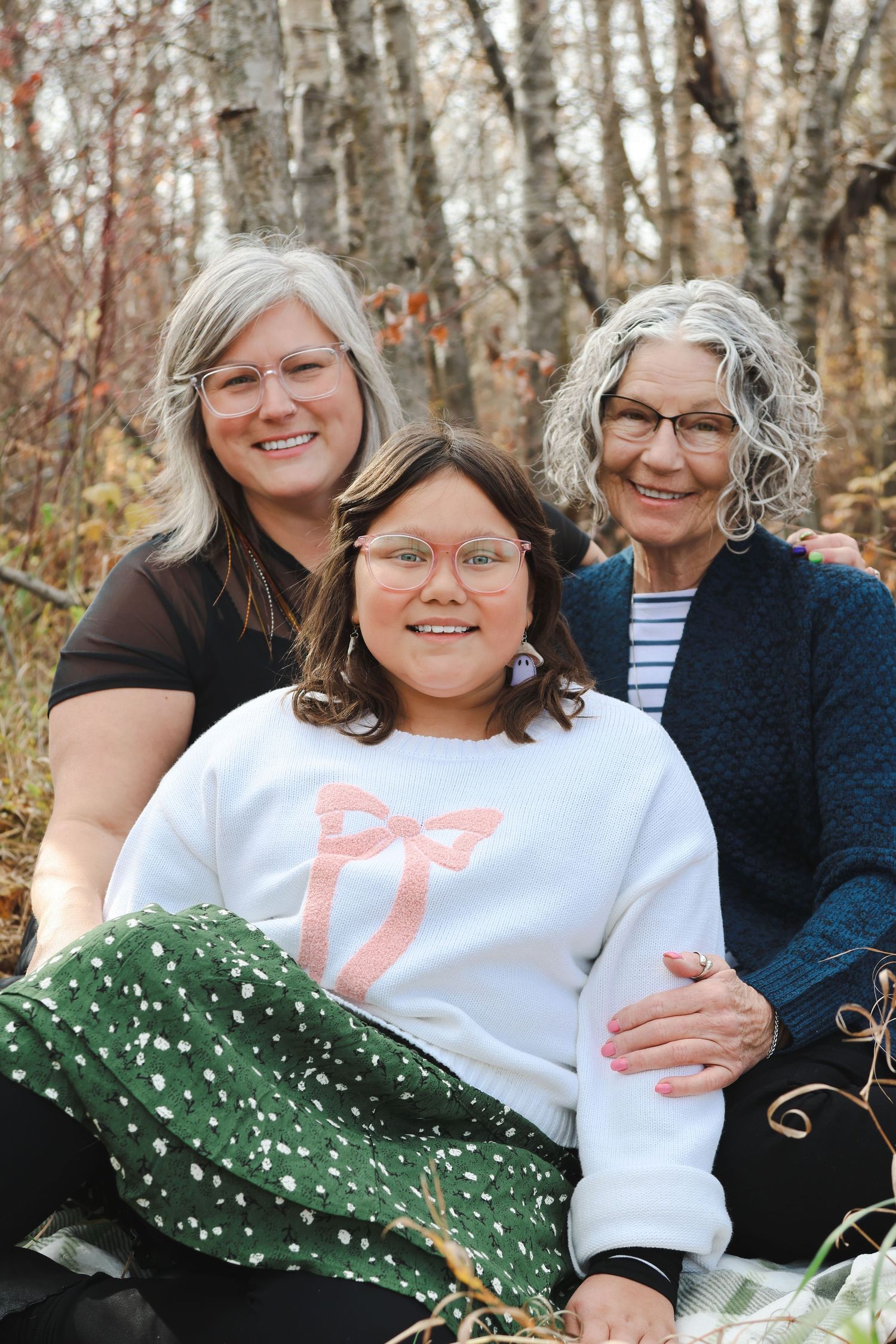 Three generations of women smile together outdoors in autumn, wearing glasses and casual clothing among fall foliage.