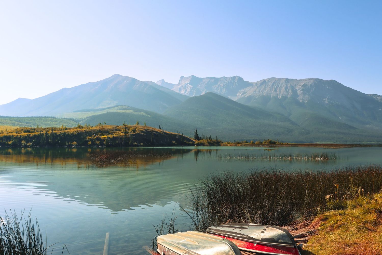 Serene mountain lake with calm reflective water, two boats on shore, autumn foliage, and majestic peaks under clear blue sky.