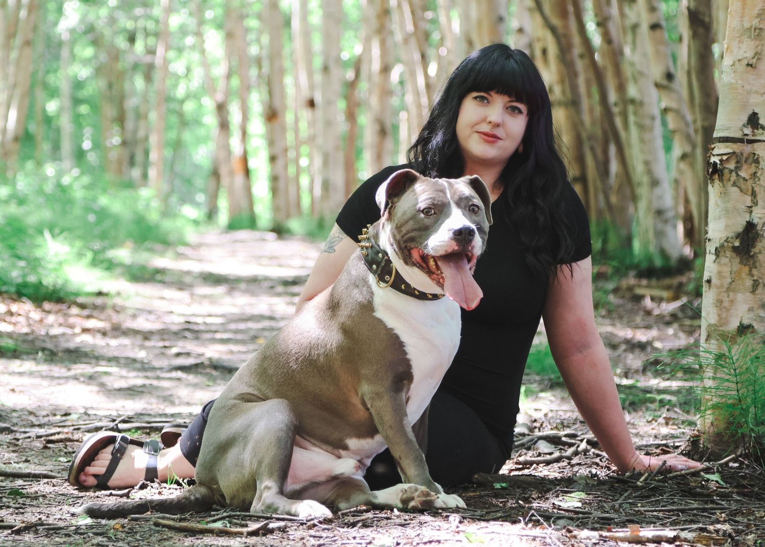 Woman with black hair sitting beside a large pit bull dog in a wooded forest path.
