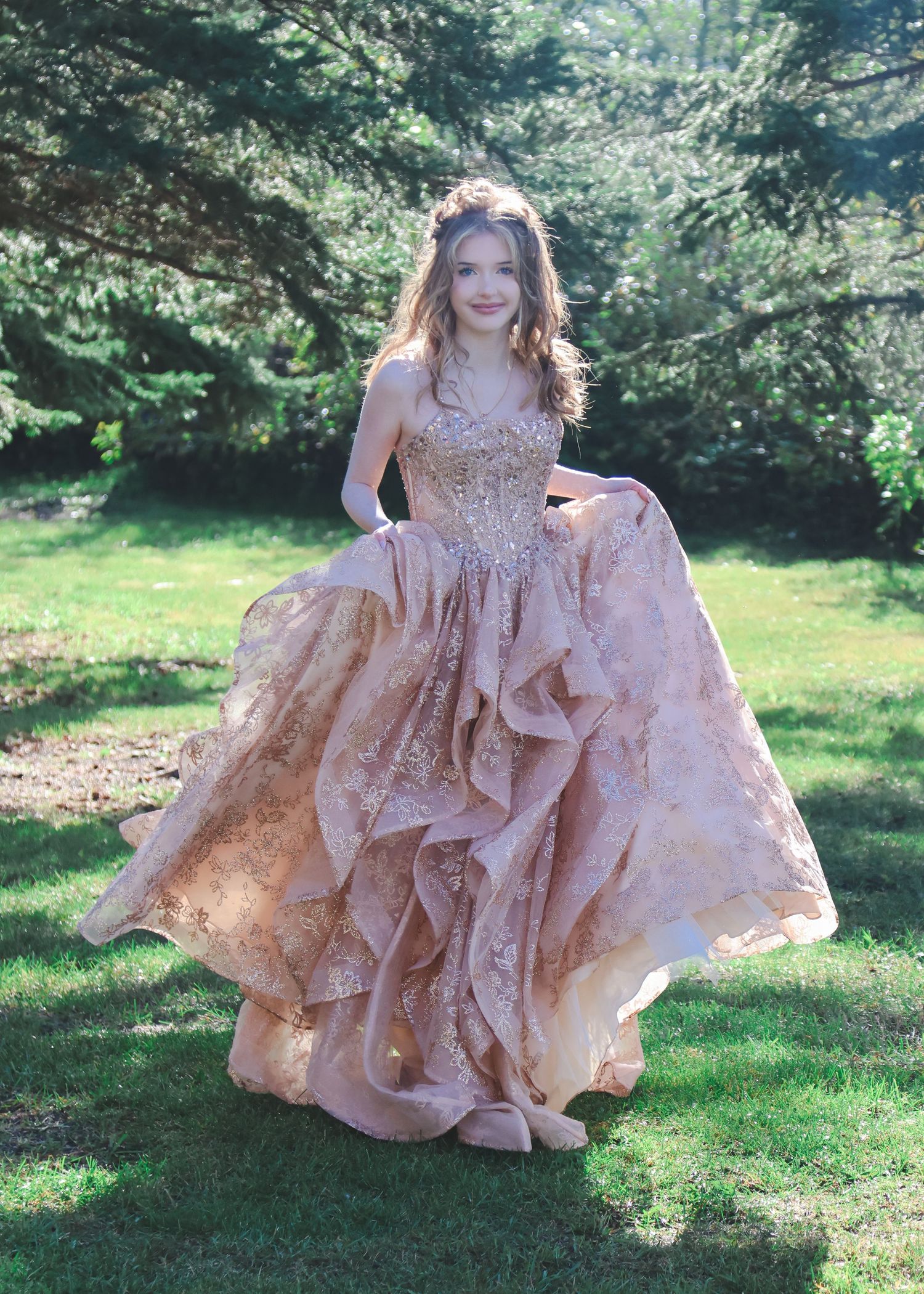 Young woman in a stunning rose gold ballgown with lace details poses outdoors in a lush green garden setting.