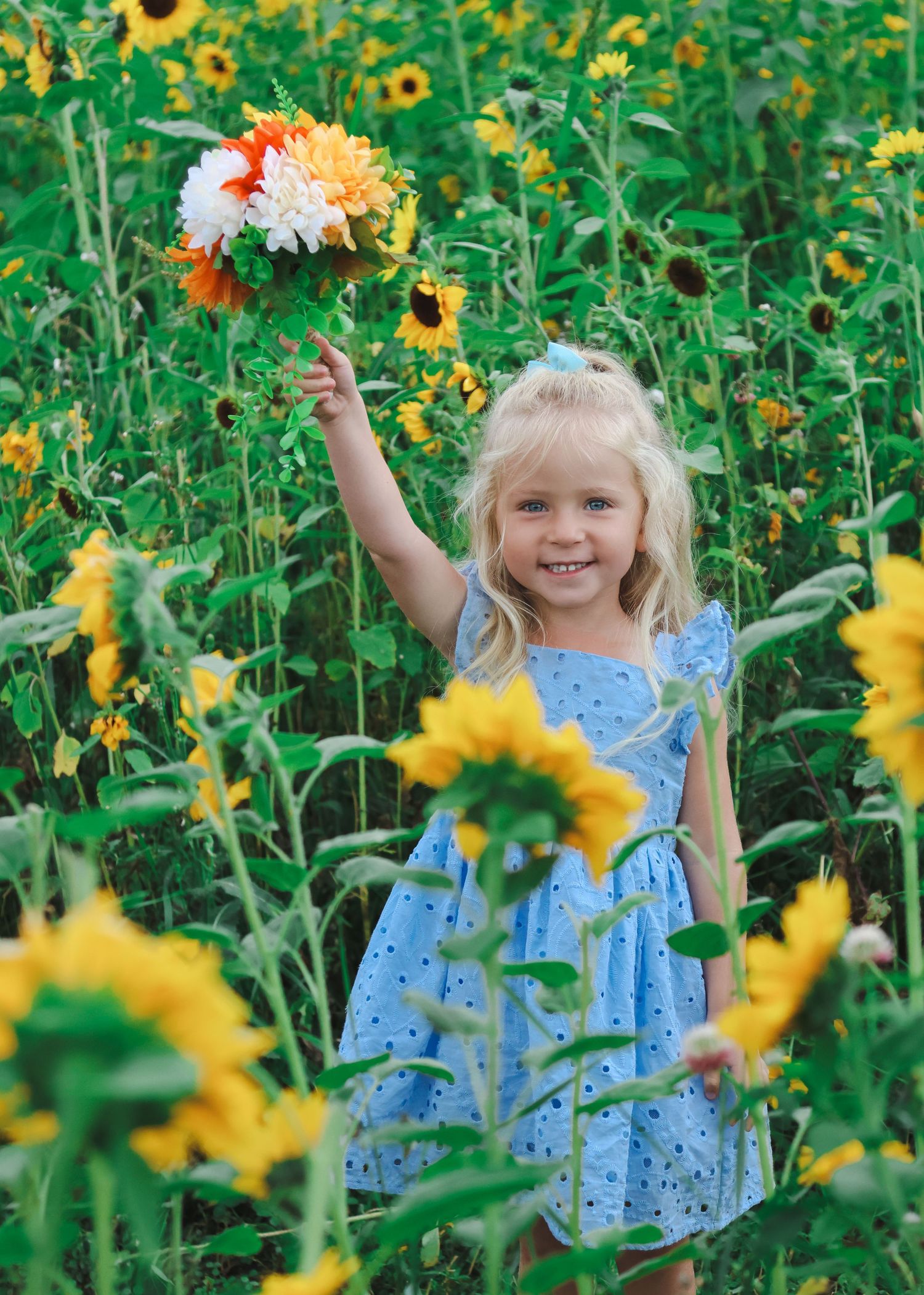 Young blonde girl in blue polka dot dress holding colorful bouquet while standing in sunny sunflower field.