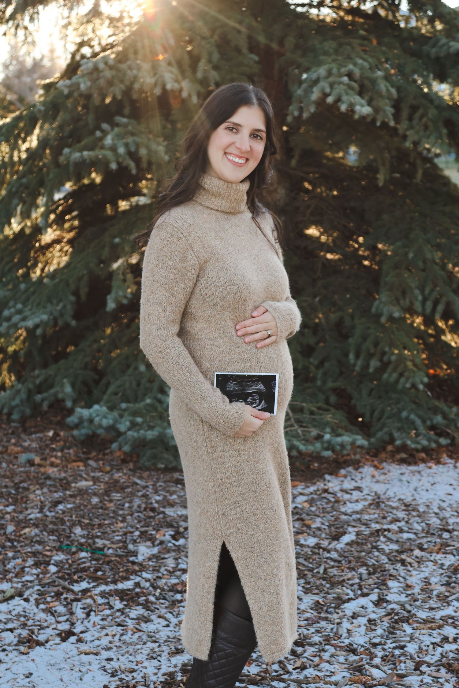 Pregnant woman in beige turtleneck dress holding ultrasound photo outdoors in winter snow.