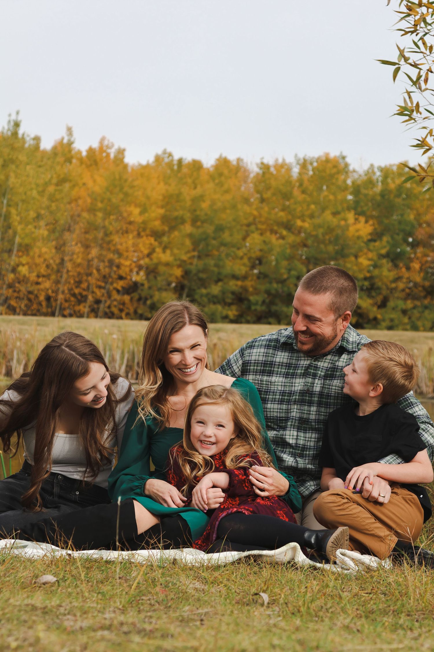 Happy family of five sits together on grass laughing with golden autumn trees in background.