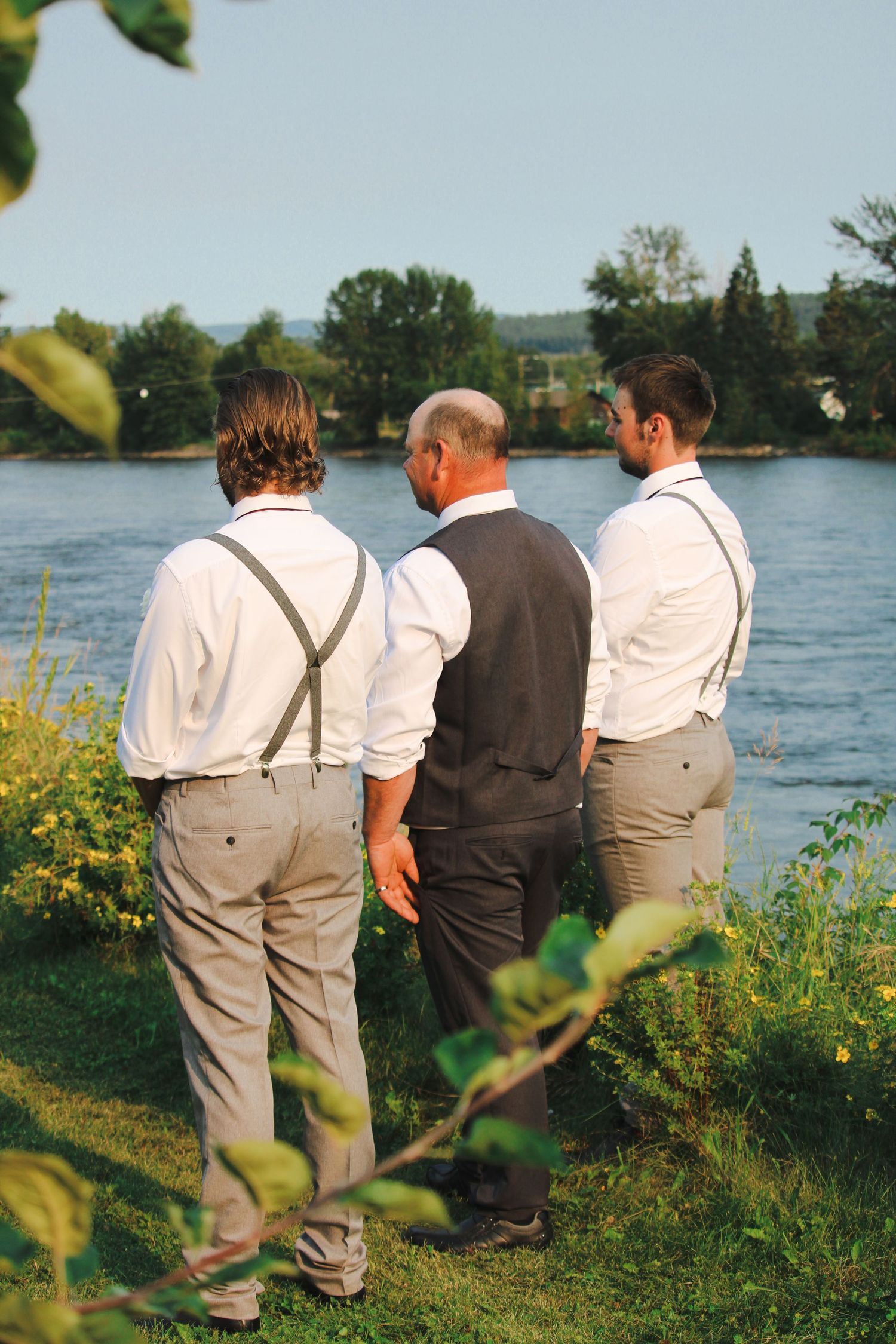 Three men in white shirts and suspenders stand together overlooking a serene river surrounded by lush greenery.