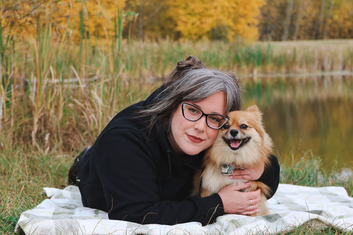 Woman with gray hair and glasses poses with a Pomeranian dog by a lake in autumn.