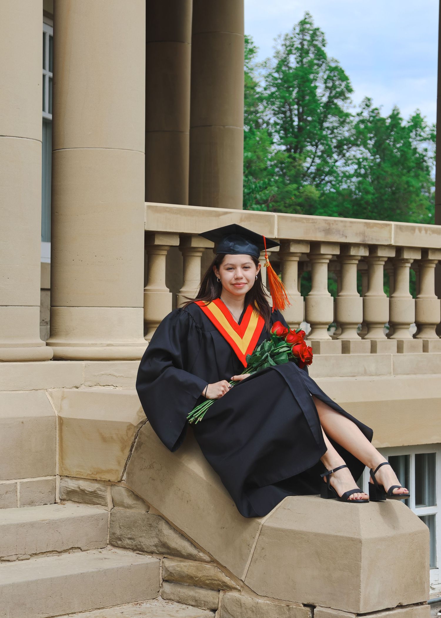 Graduate in black cap and gown holding red roses seated on stone steps.