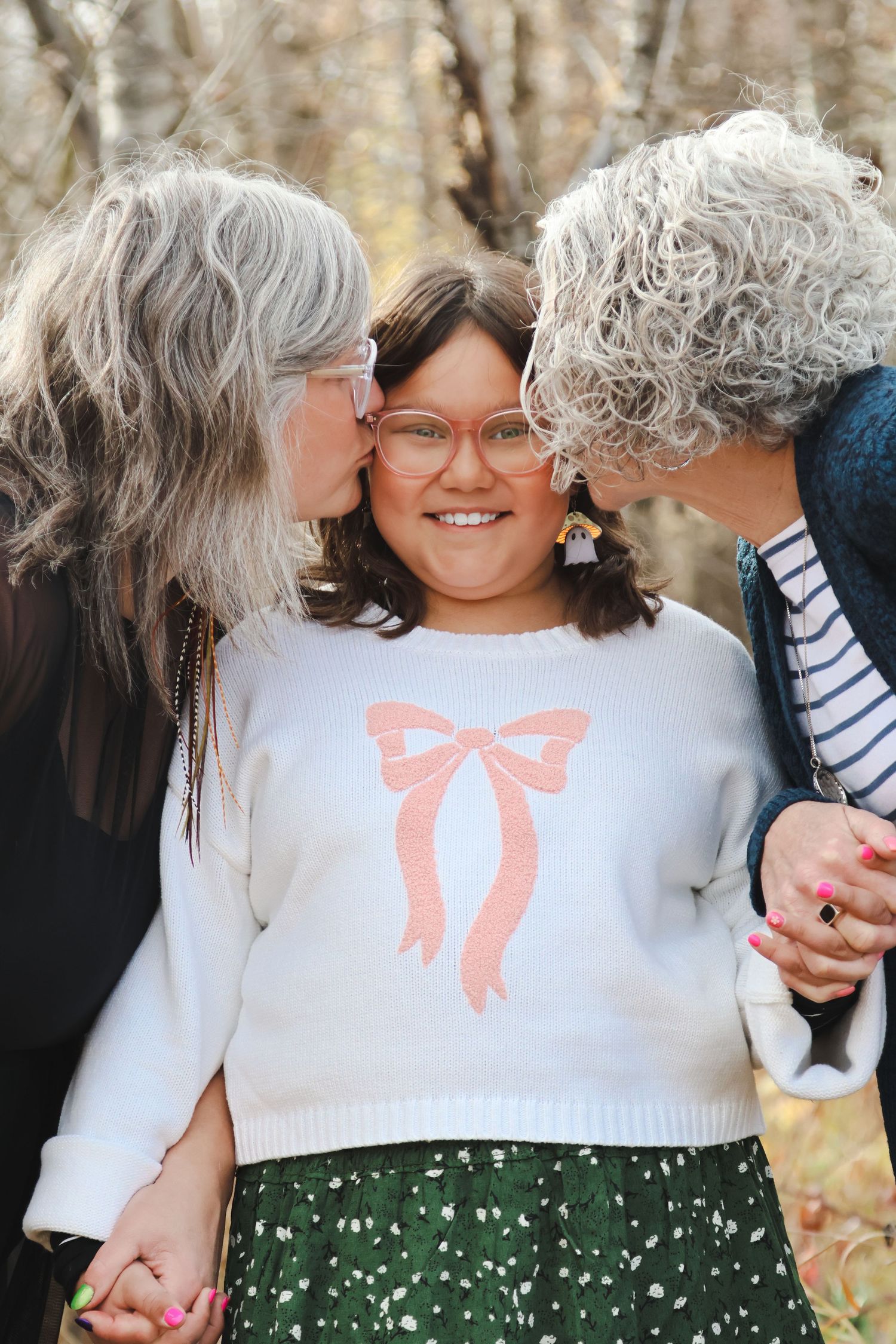 Two older women with gray hair kissing the cheeks of a smiling young girl wearing a bow sweater outdoors.