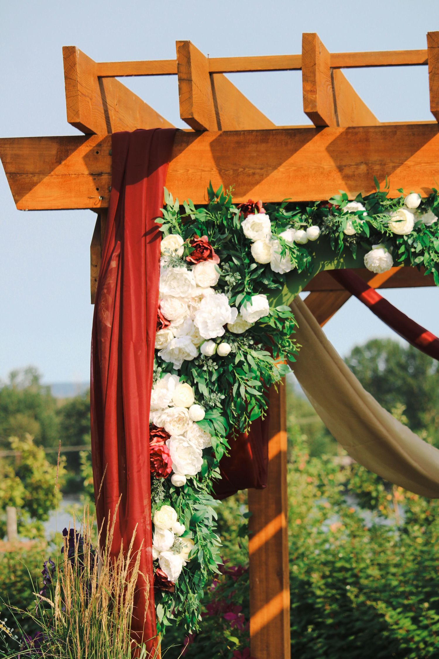 Wooden pergola decorated with white and red floral garland and red draped fabric for an outdoor wedding ceremony.