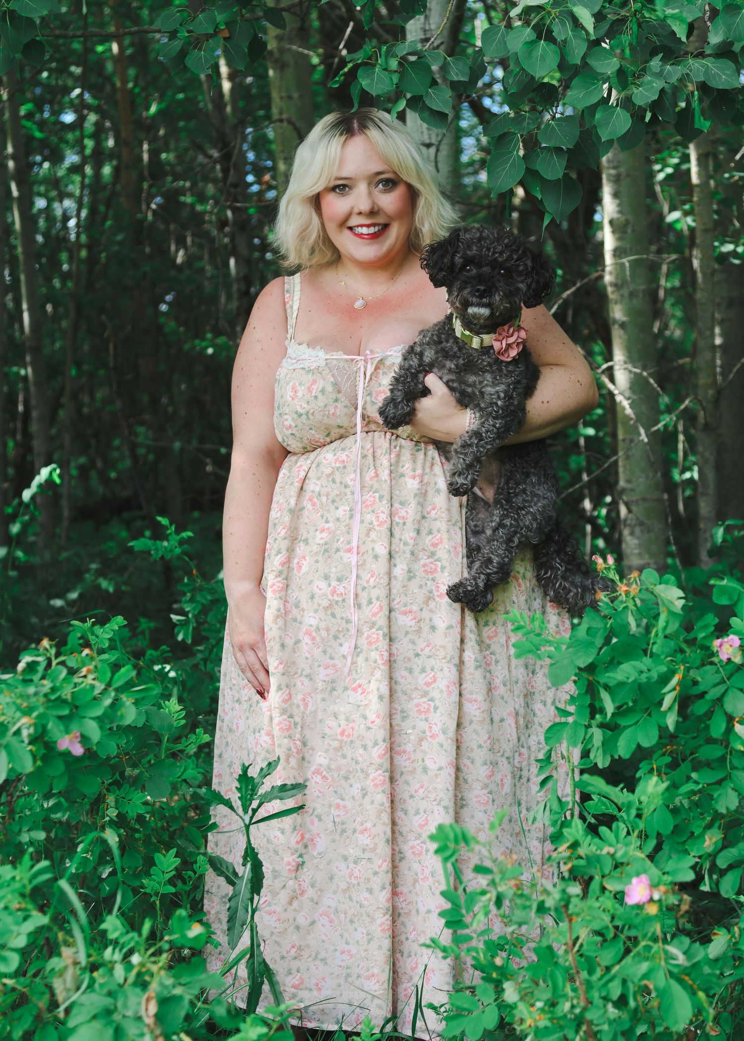 Woman in floral maxi dress holding a black poodle in a lush green forest setting.