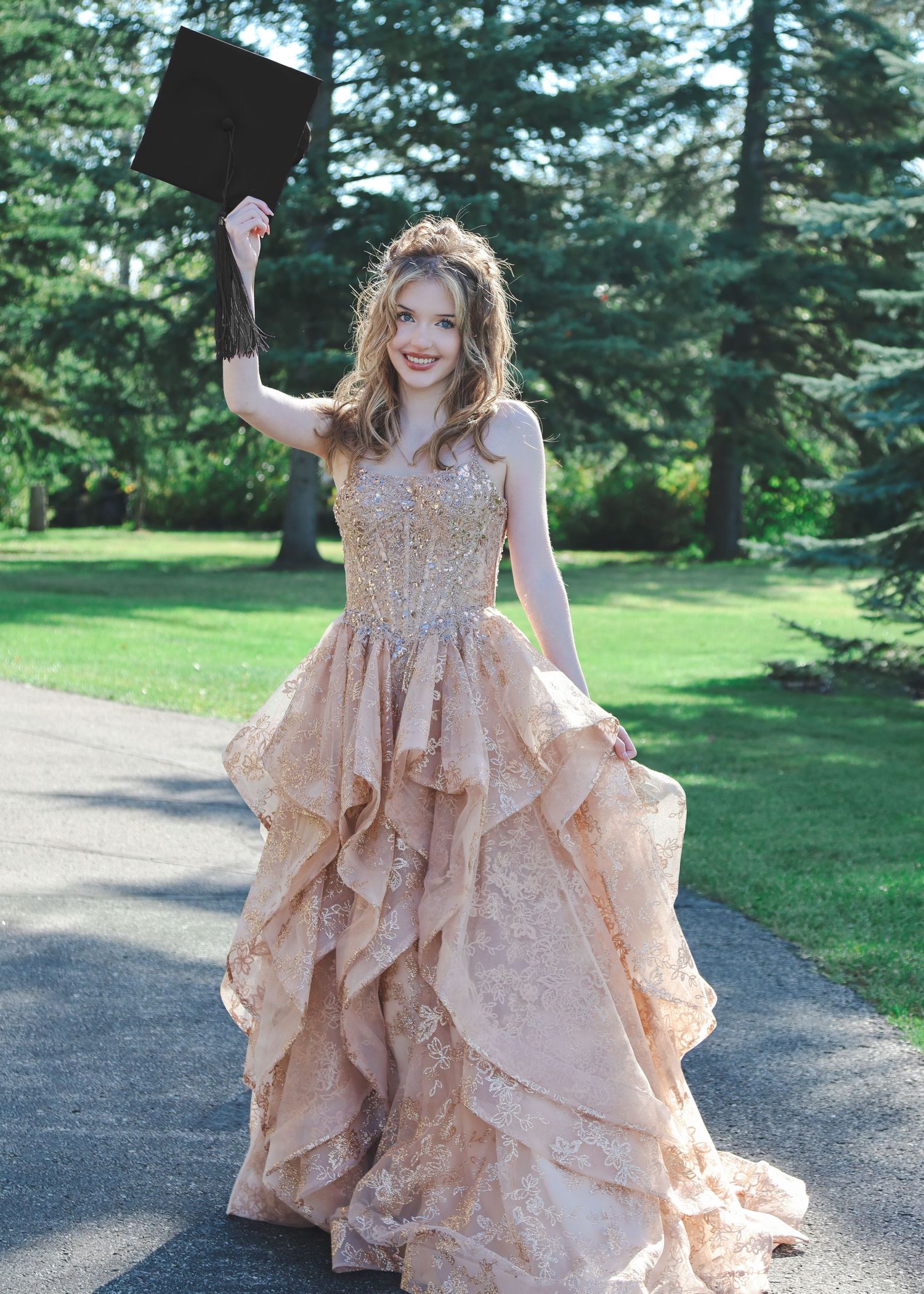 Young woman in gold lace ballgown holding graduation cap aloft, celebrating outdoors on a tree-lined path.