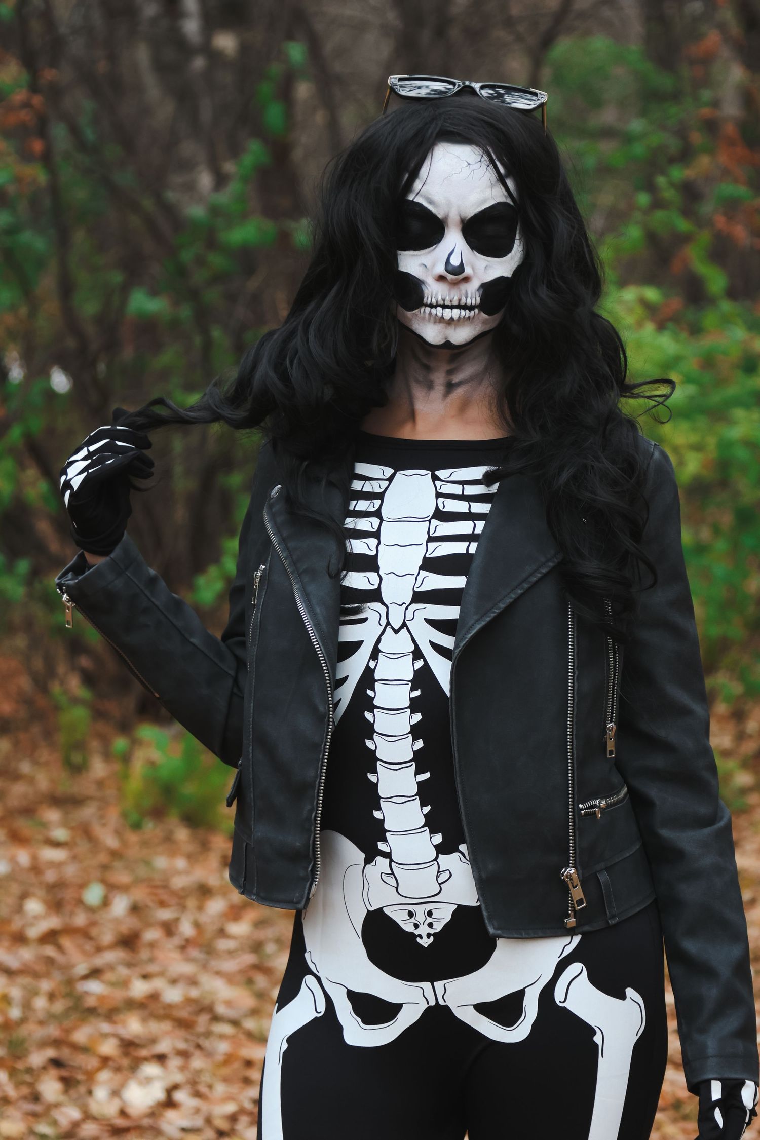 Woman in skeleton costume and skull face paint wearing black leather jacket poses in autumn forest.