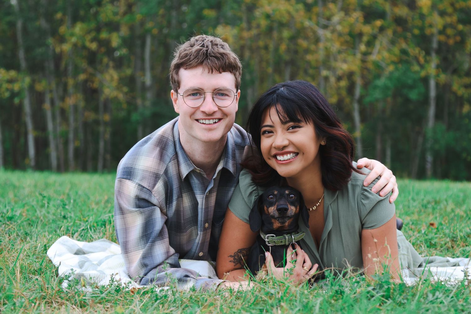 A smiling couple lying on grass holding a small black dachshund puppy with trees in the background.